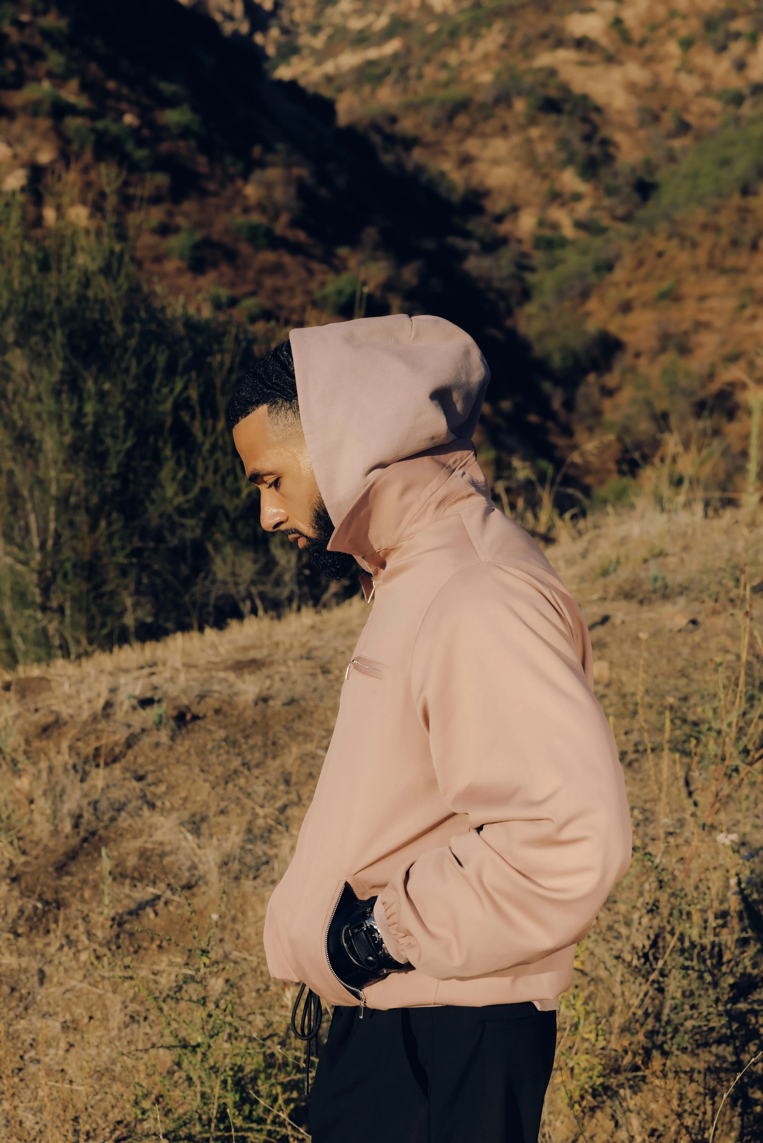 A man in a beige hoodie and black pants standing outdoors on a hillside with dry grass and shrubs, looking down with a contemplative expression.