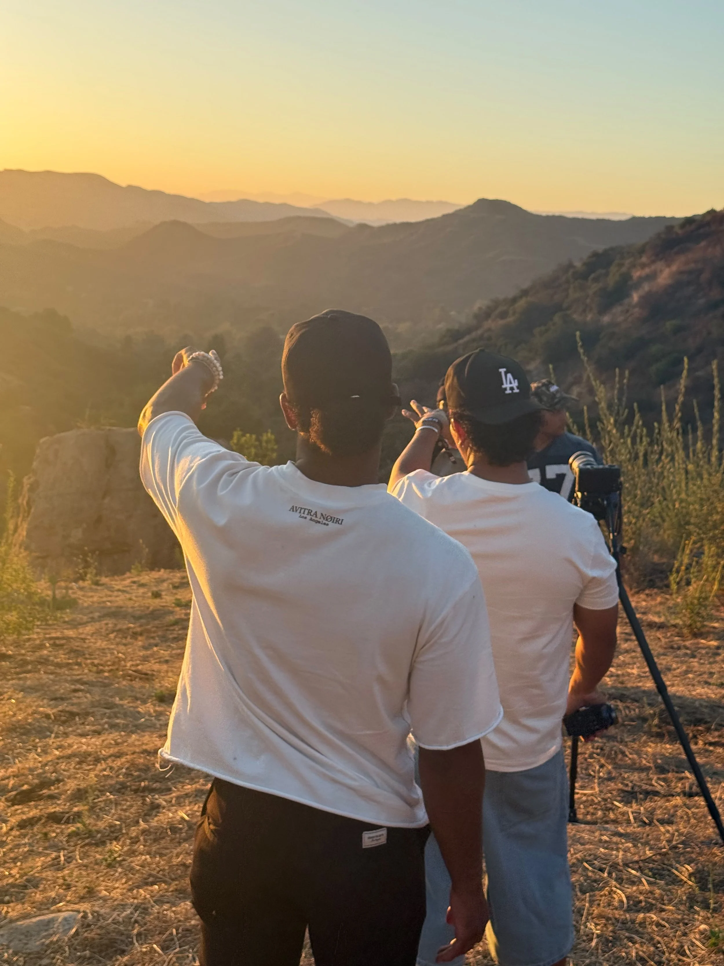 Three people standing outdoors on a hillside at sunset, with mountains in the background, one person pointing towards the horizon.