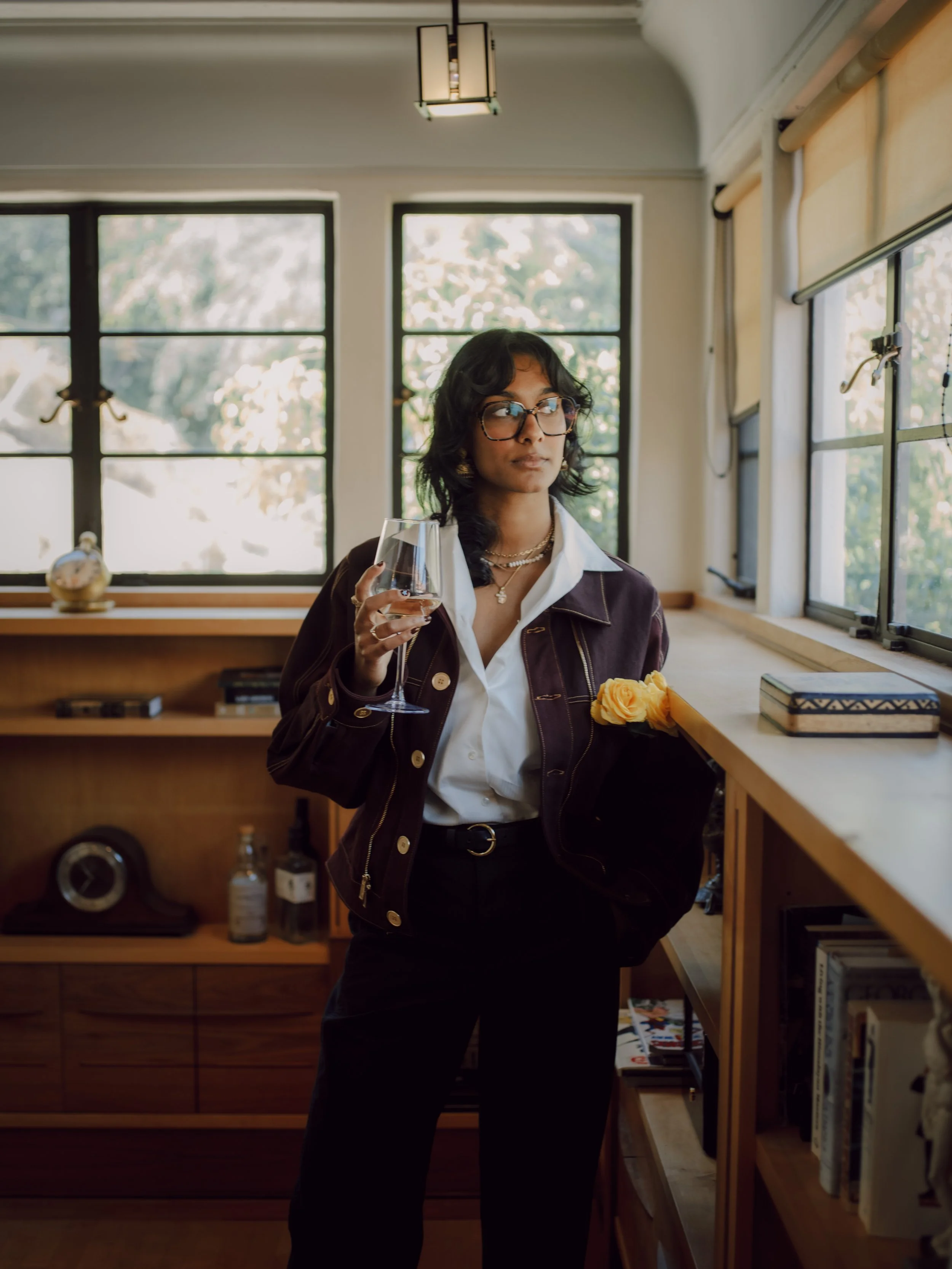 A young woman wearing glasses and a layered necklace stands indoors near a wooden bookshelf, holding a wine glass in one hand and a yellow flower on the bookshelf. She has shoulder-length dark hair and is dressed in a white shirt and dark jacket. The