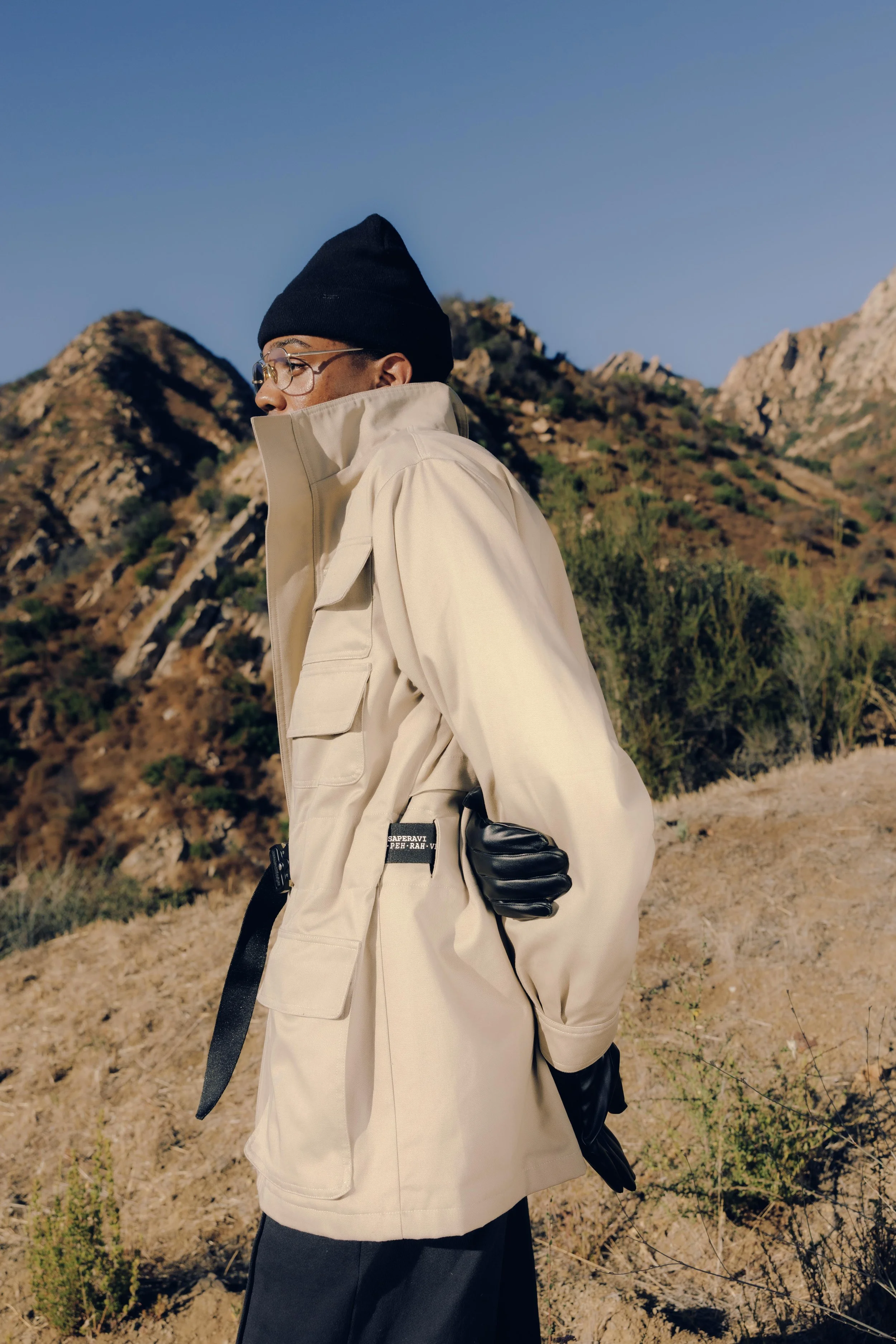 A person dressed in a beige trench coat, black beanie, and glasses walking outdoors in a mountainous area with dry grass and sparse greenery under a clear blue sky.