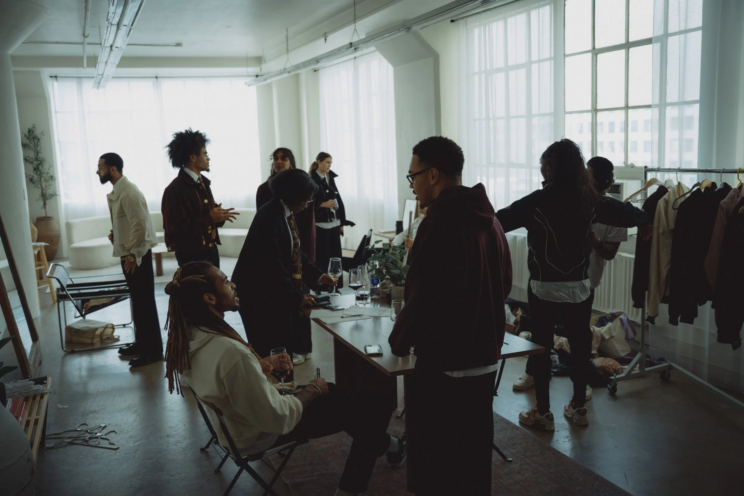 People gathered in a bright room, some sitting, some standing, with clothing racks and a table, possibly preparing for a fashion event or photo shoot.