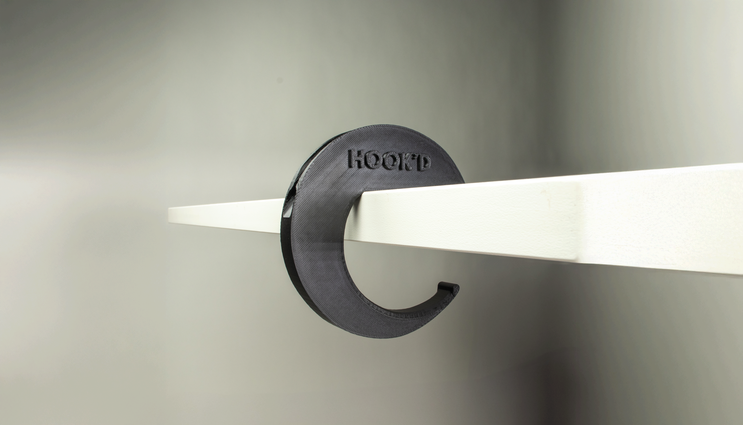 Close-up of a black, C-shaped hook labeled 'HOOKD' attached to a white shelf in a plain, gray room.