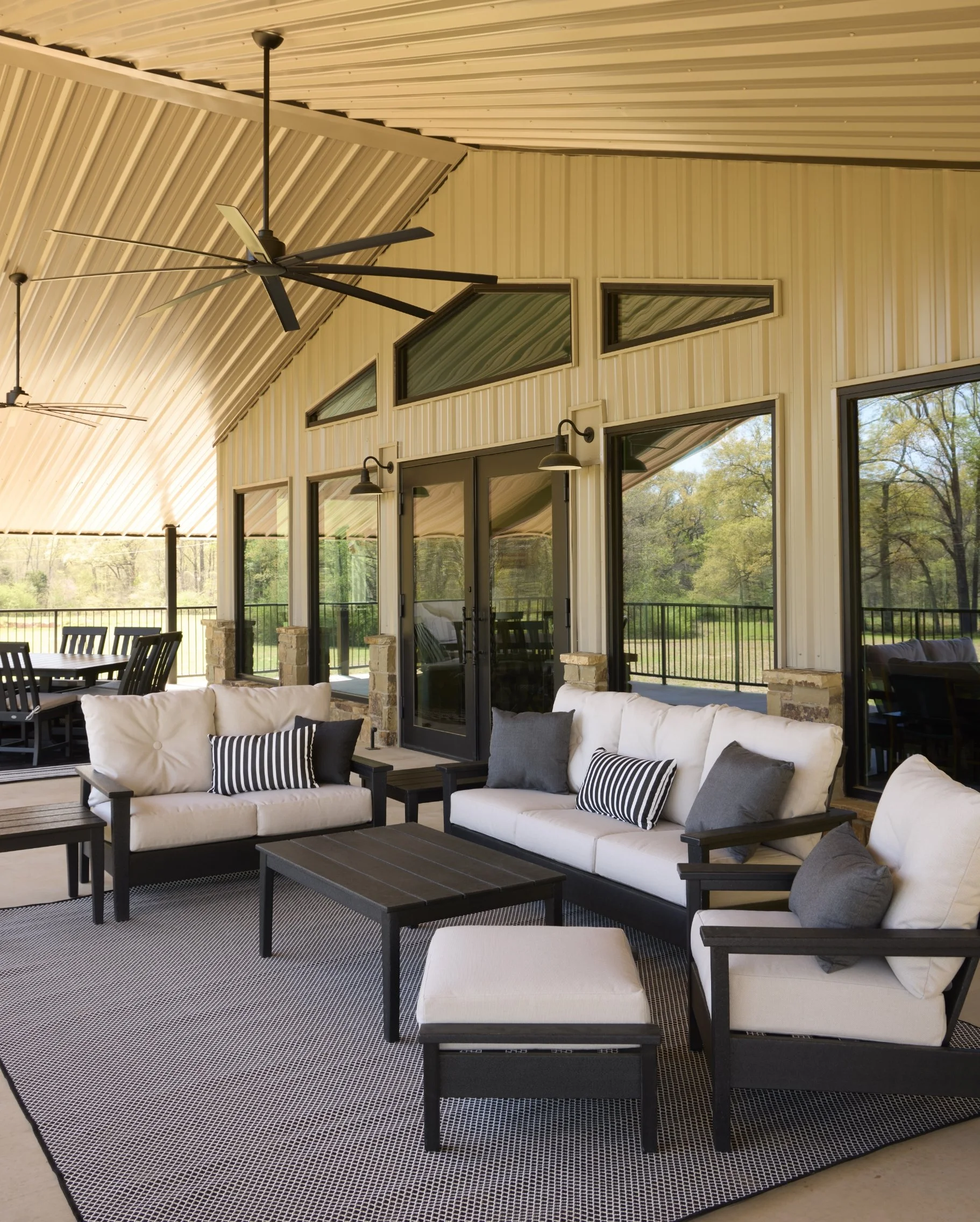 Covered outdoor patio with beige sofas, black and white striped pillows, black coffee table, ceiling fans, and large windows overlooking greenery.