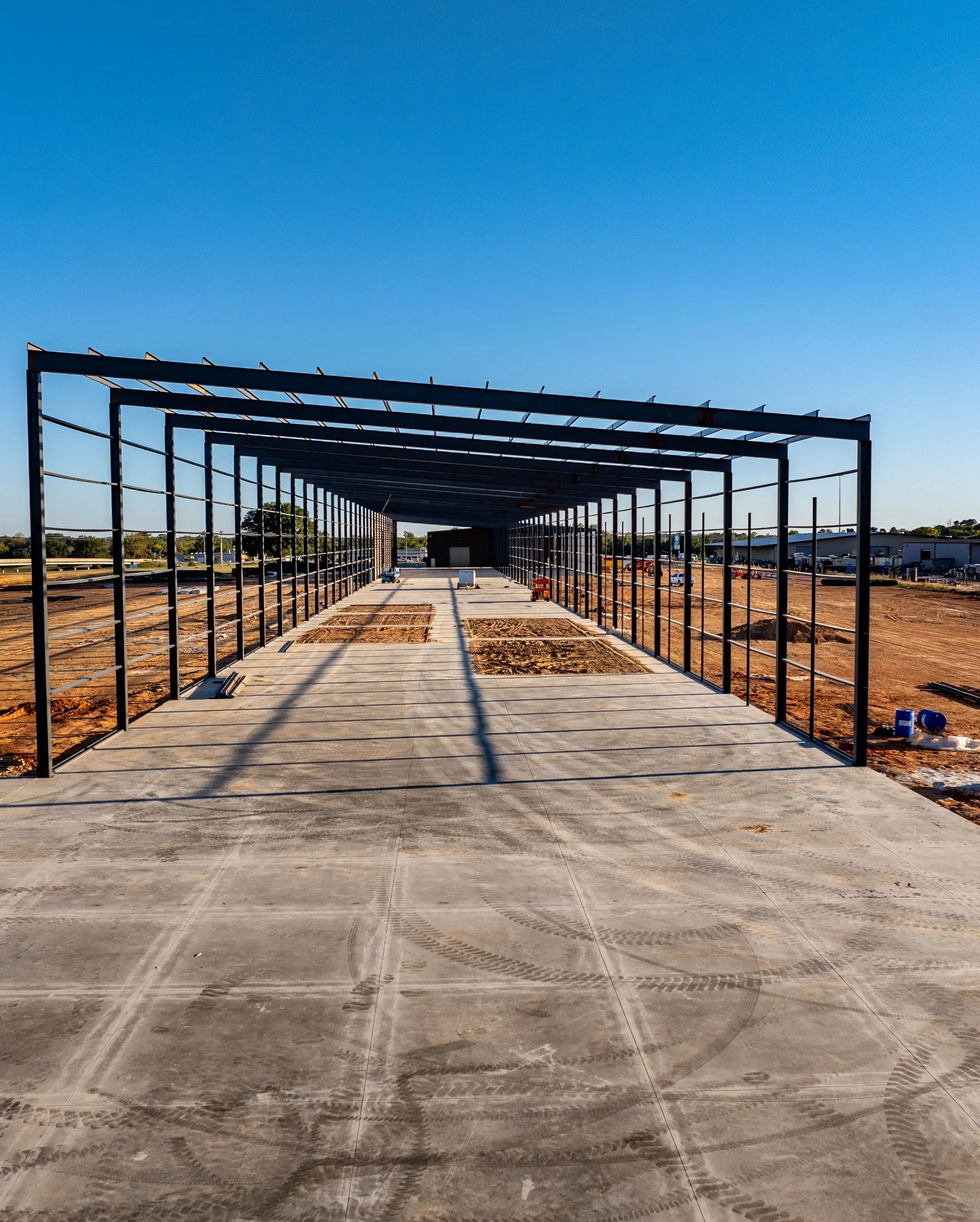 Construction site with a building frame on a concrete foundation, surrounded by dirt, under a clear blue sky.