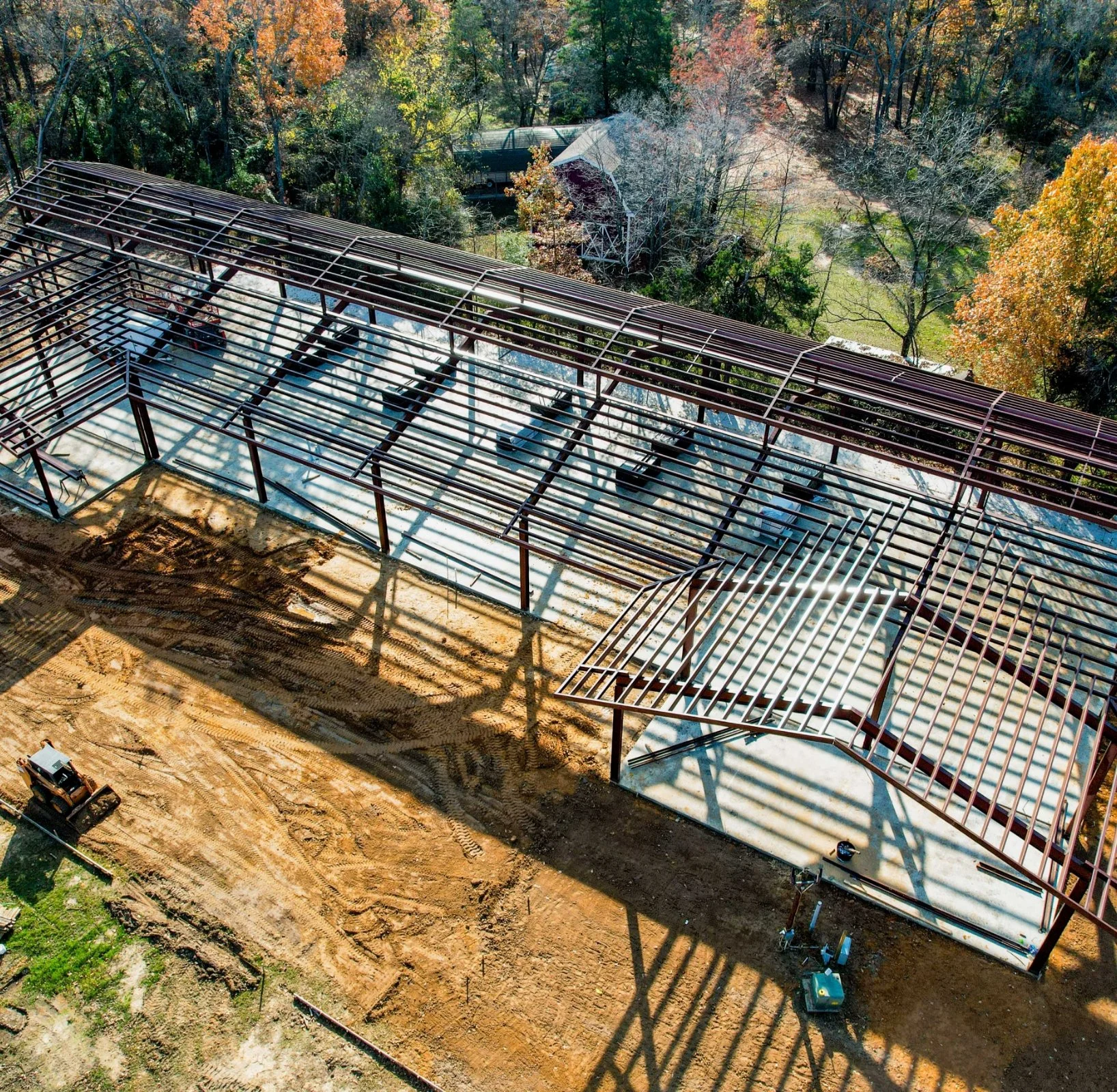 Aerial view of a building under construction with steel framework, surrounded by trees and dirt ground.