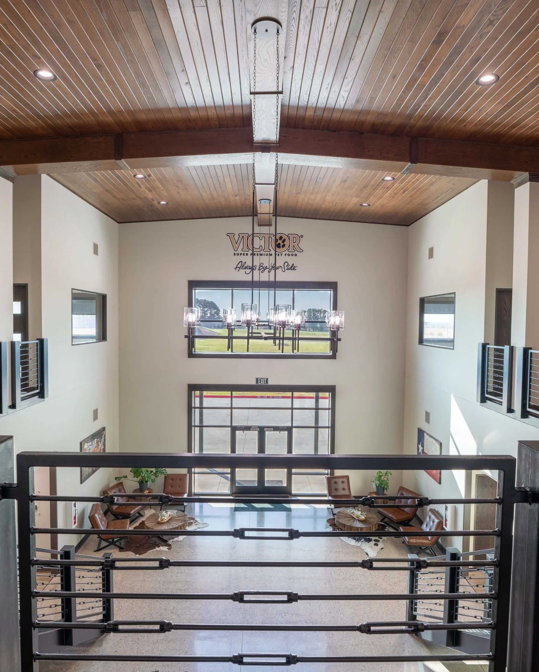 Interior view of a spacious building with a wooden ceiling, chandelier, and large windows overlooking a field, featuring a seating area with chairs and tables near the entrance.