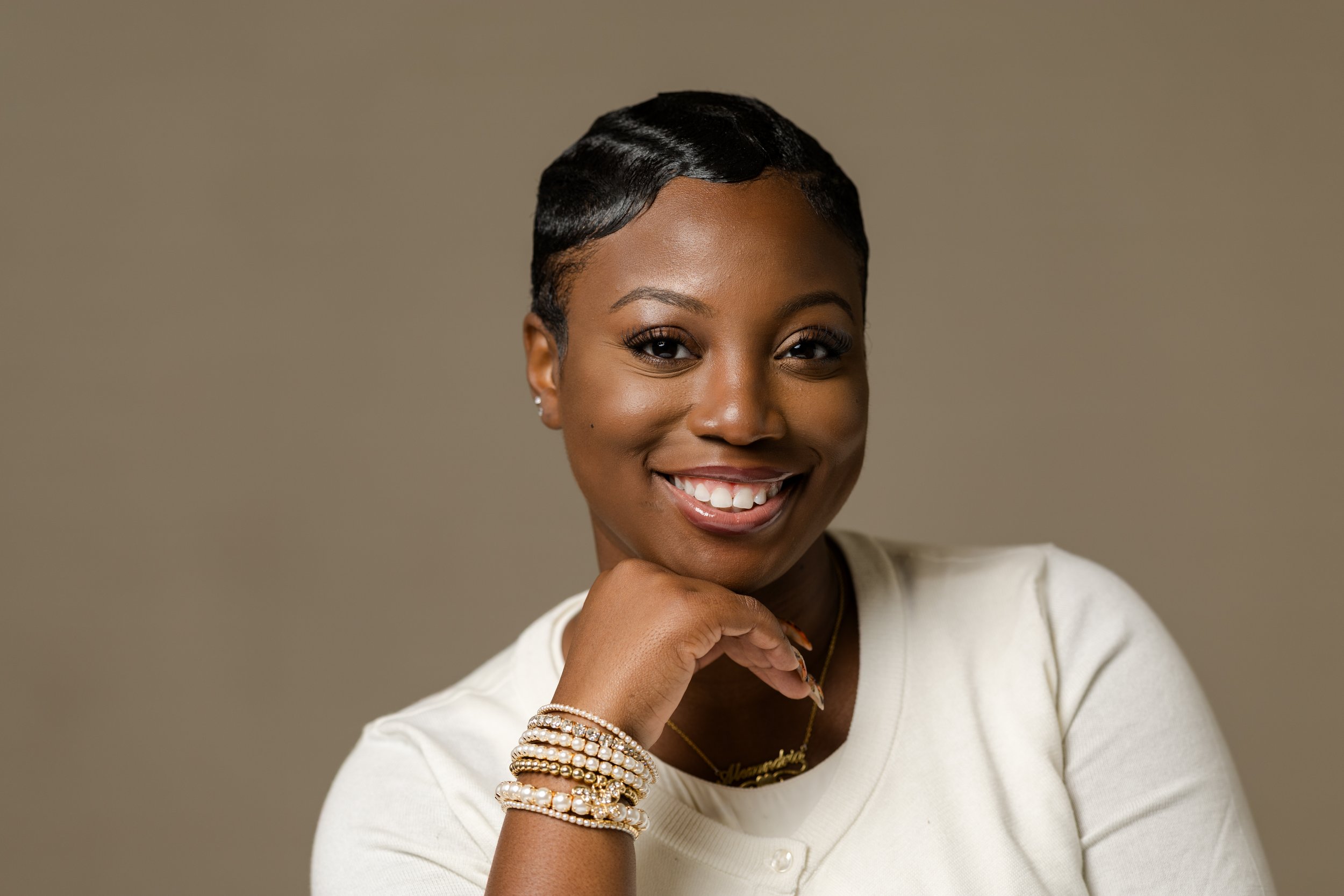 Portrait of a smiling Black woman with short, wavy hair, wearing pearl bracelets and a white top against a neutral background.