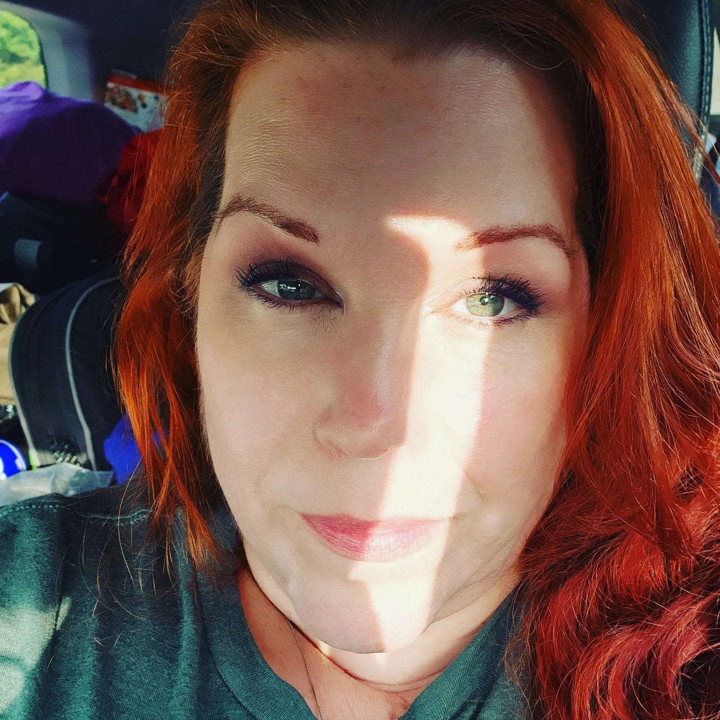 Close-up portrait of a woman with red hair and green eyes, with sunlight casting a shadow on part of her face, sitting inside a vehicle.