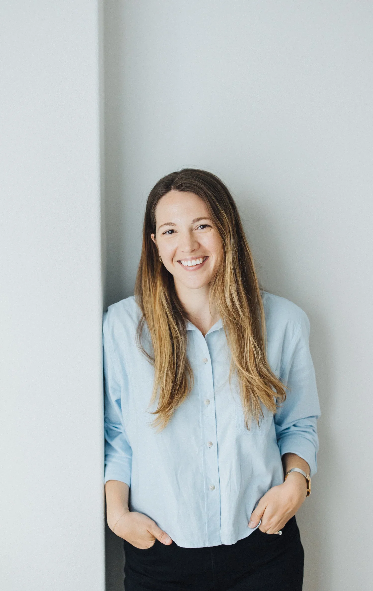 A woman with long, light brown hair, wearing a light blue button-up shirt and black pants, standing against a light gray wall, smiling with her hands in her pockets.