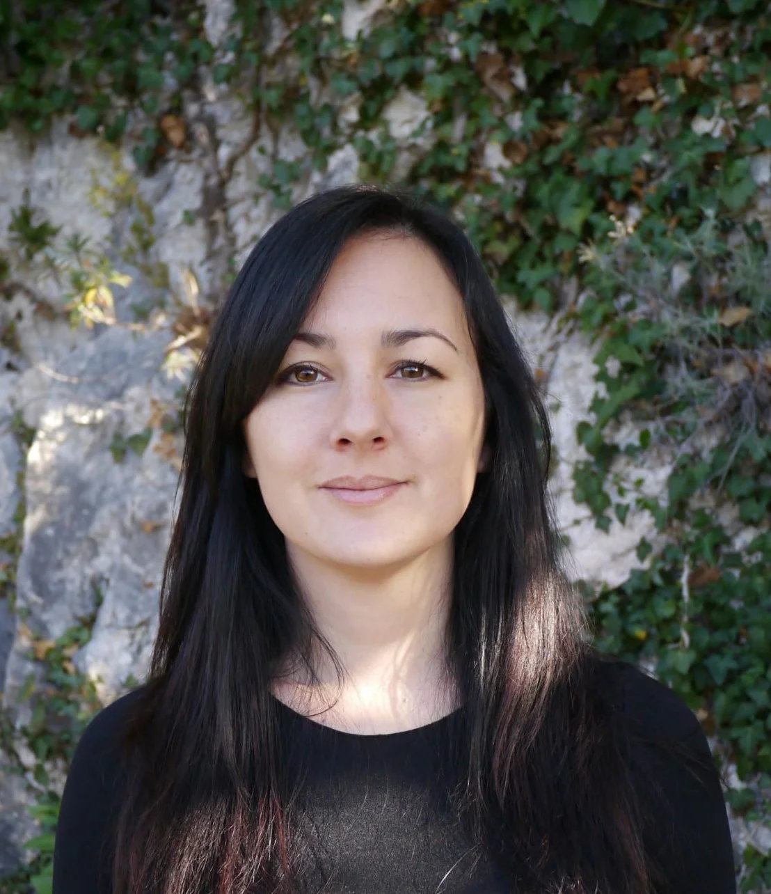 A woman with long dark hair smiling outdoors with greenery and a rock wall in the background.