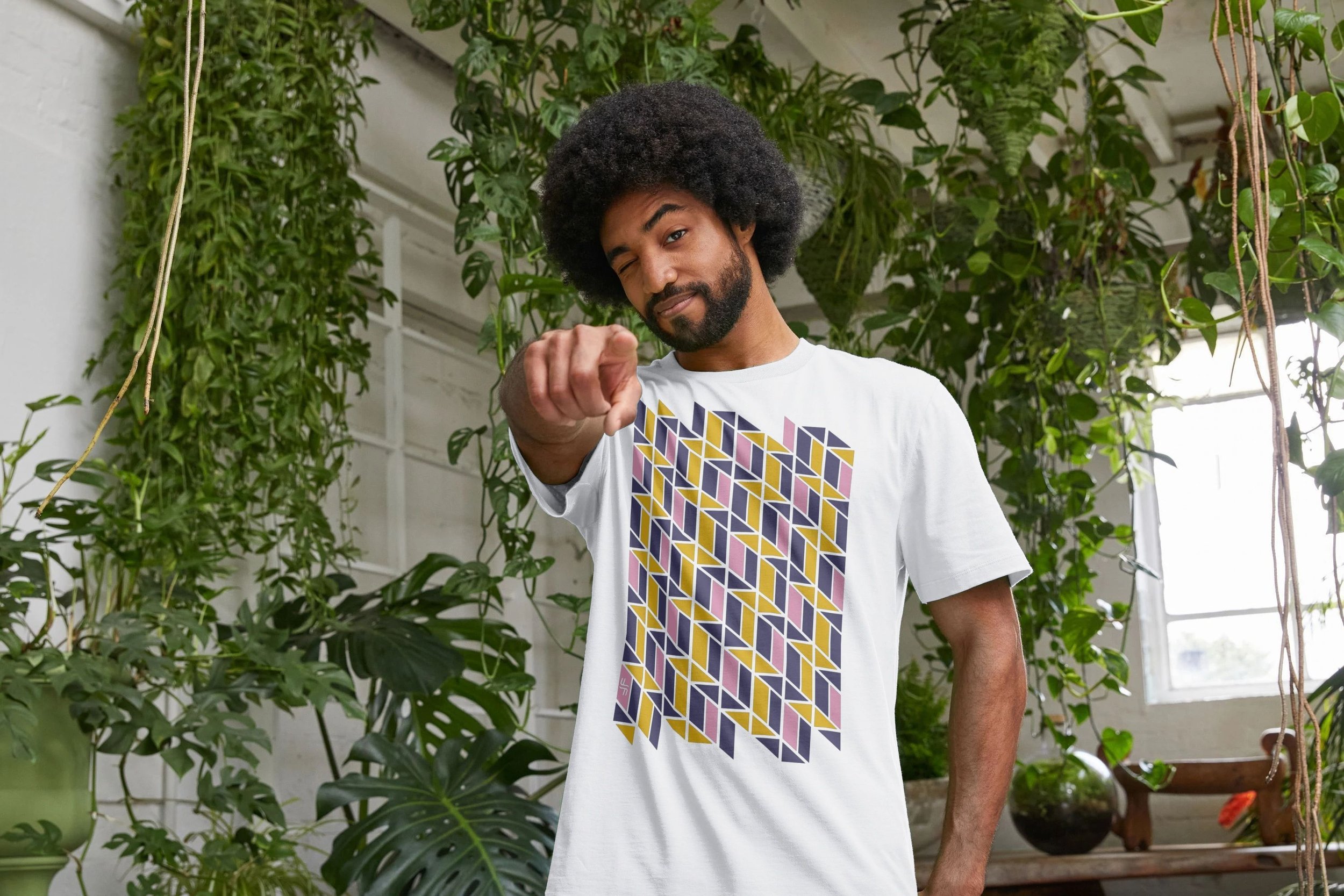 A man with a curly afro and beard pointing at the camera in a greenhouse filled with plants.