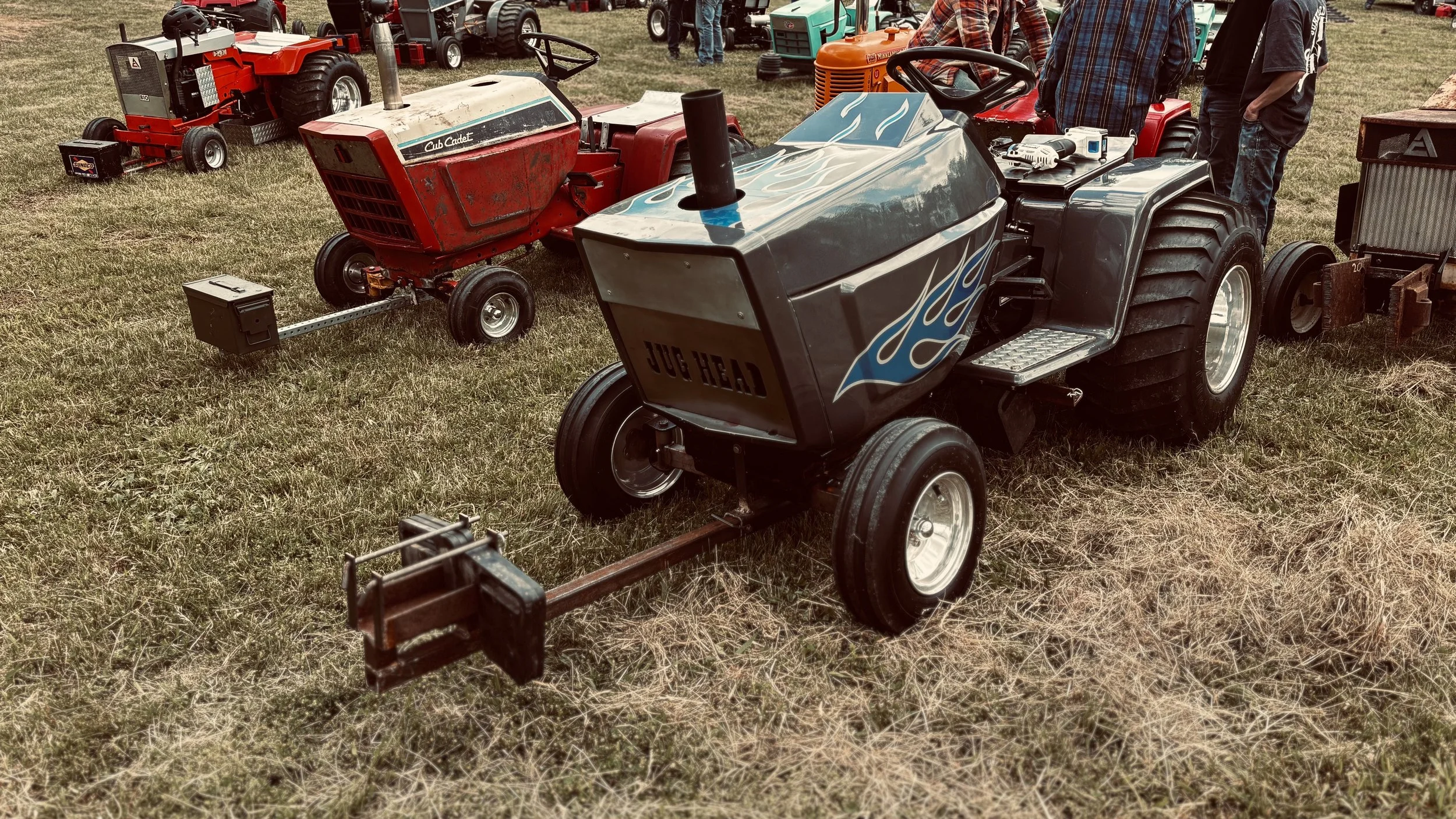 A small red tractor and a larger gray tractor with blue flame decals displayed on a grassy field at a tractor show, with several people and other tractors in the background.