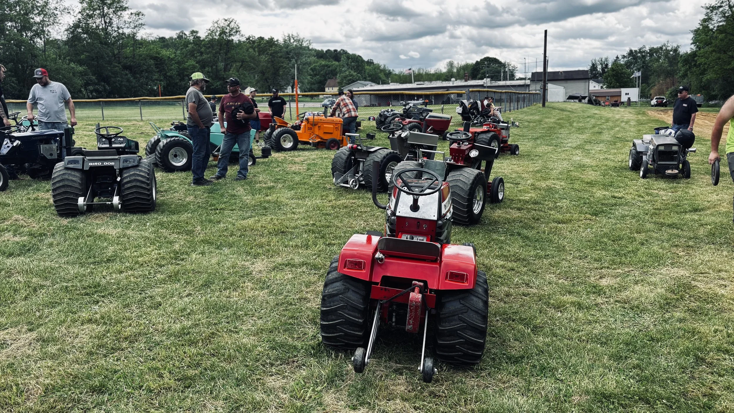 Various small lawn tractors and mini-plows displayed on a grassy field at an outdoor event, with people walking and examining the equipment under cloudy skies.