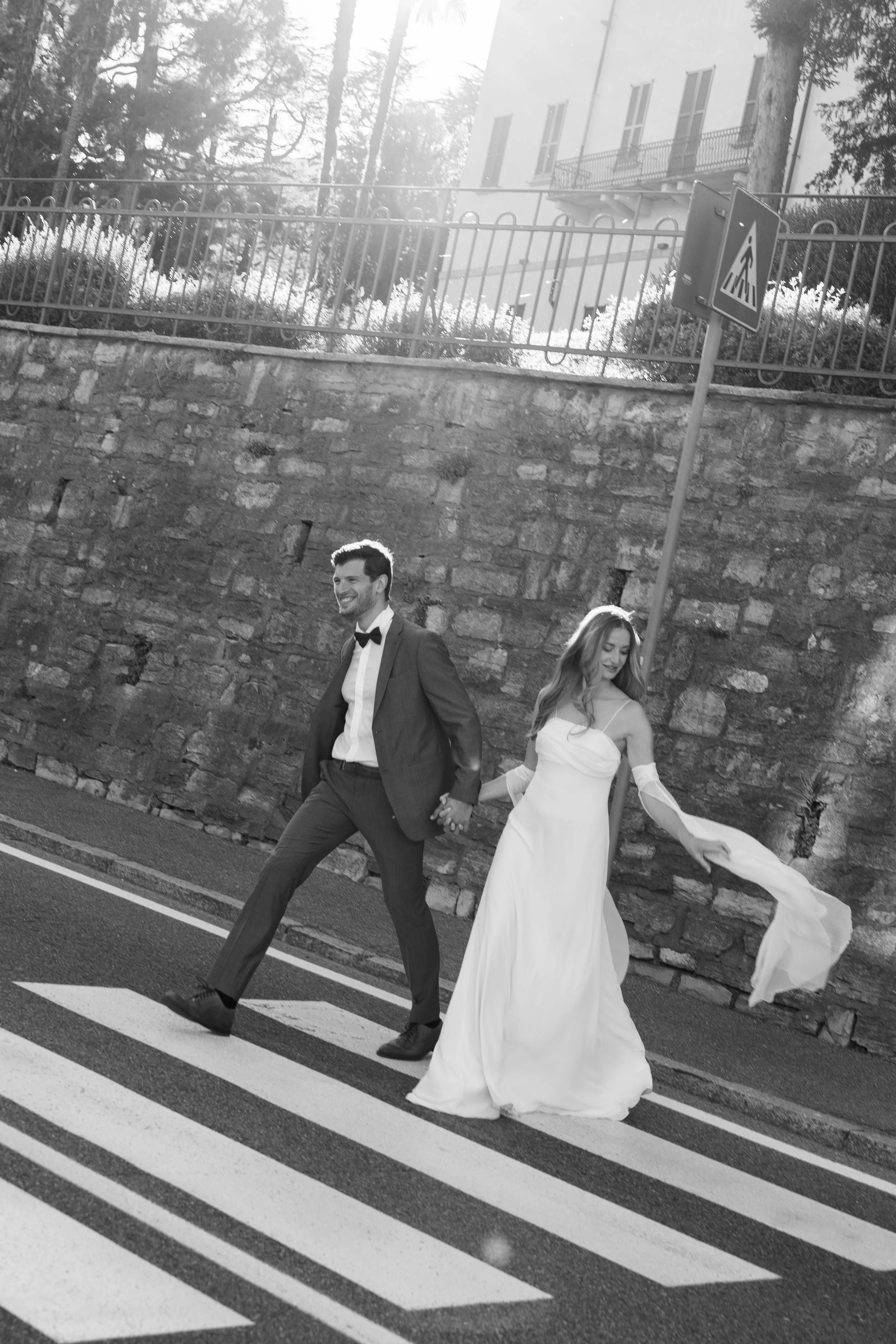 A couple dressed in wedding attire walking hand in hand across a crosswalk outdoors, with a stone wall and building in the background, in black and white.