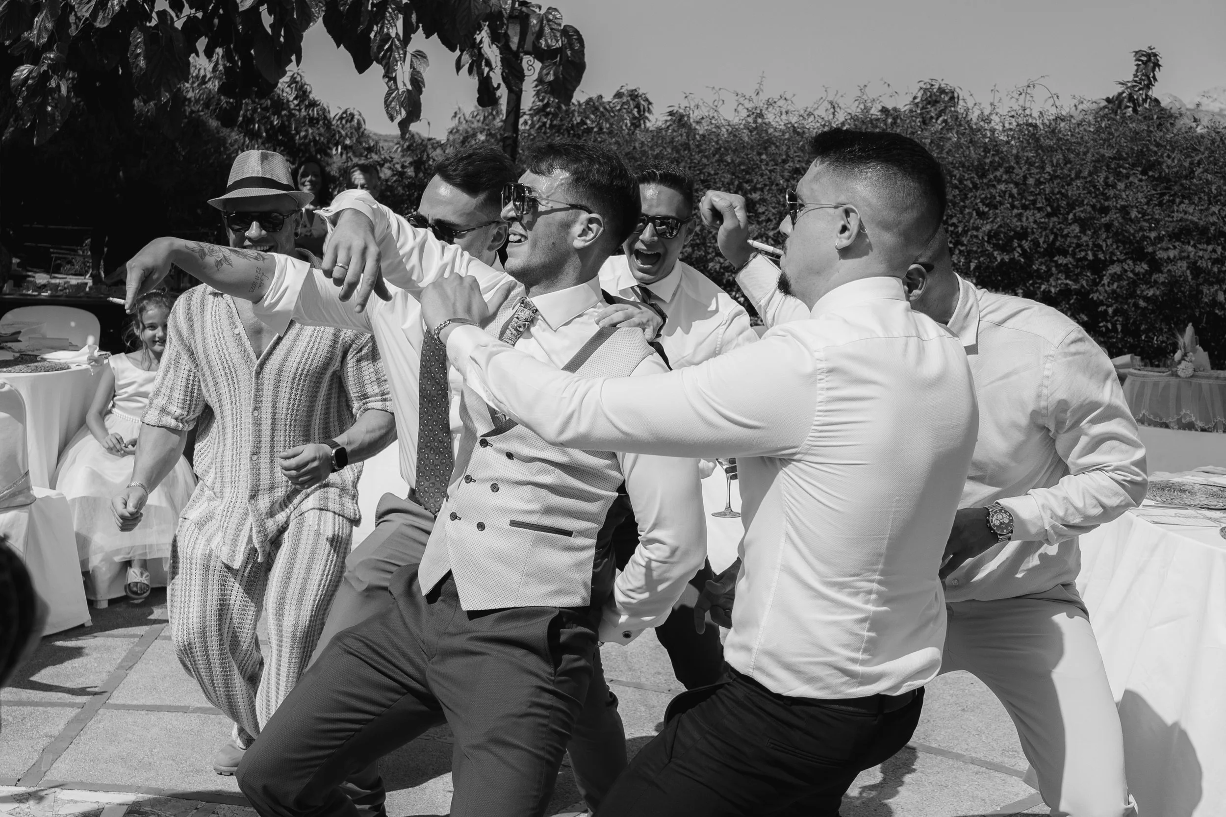 A group of men dancing and celebrating at an outdoor event, possibly a wedding, with some wearing sunglasses and suits, and a woman and children in the background
