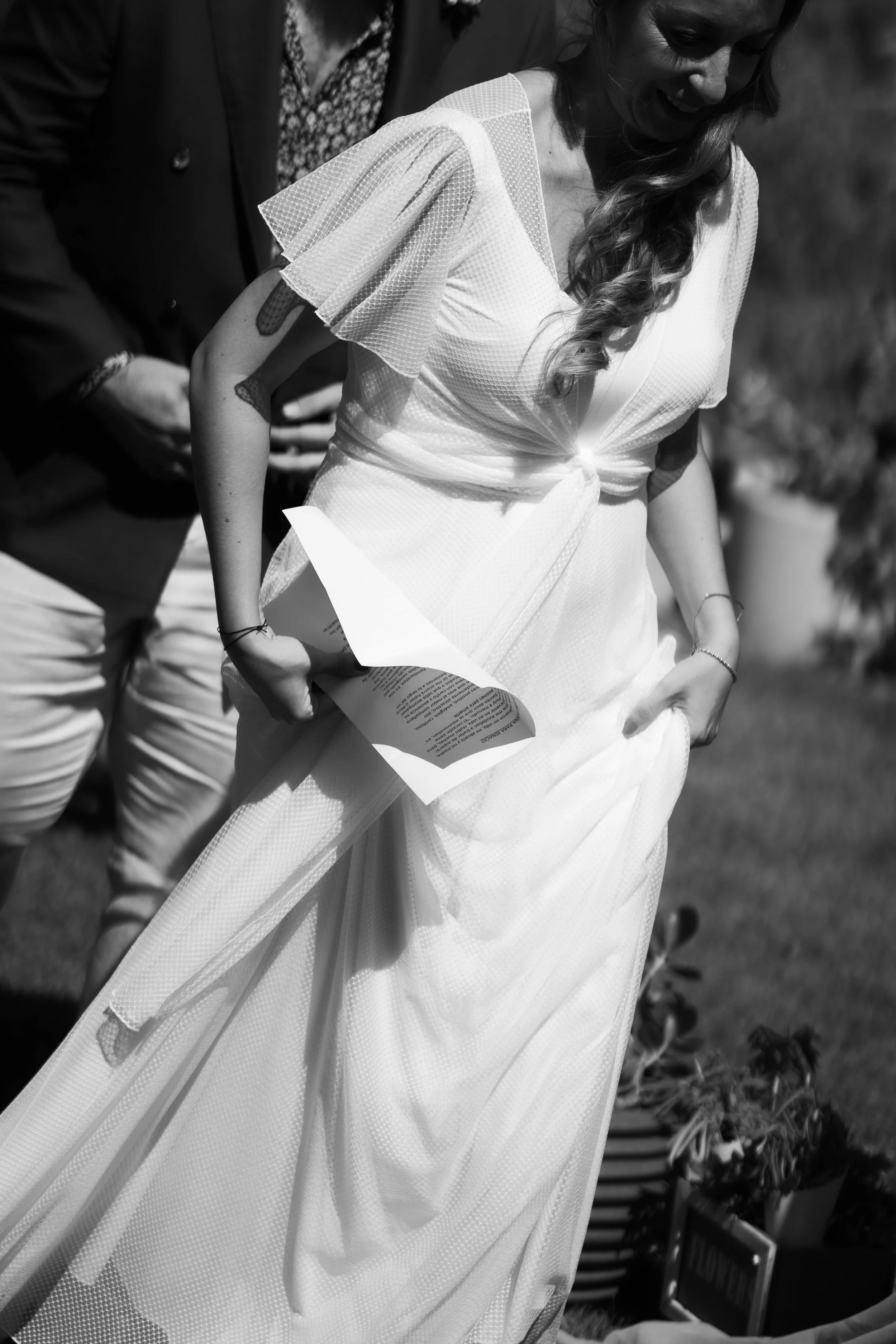 A woman in a white dress holding a program, smiling, with another person partly visible beside her, outdoors during daytime.