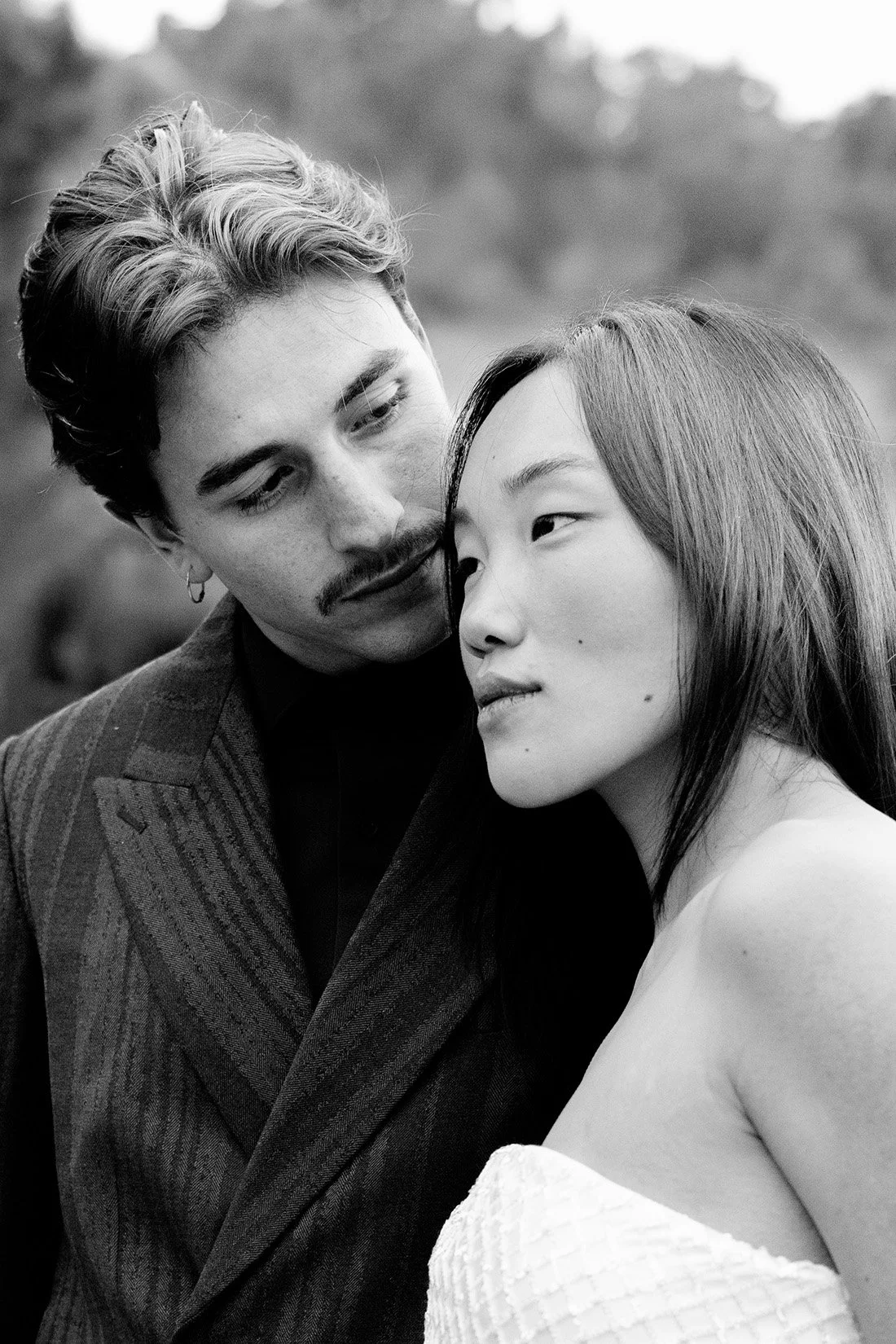 Una pareja en un primer plano, con el hombre con bigote y pelo rizado, y la mujer con cabello liso y ojos cerrados, en un entorno al aire libre en blanco y negro.