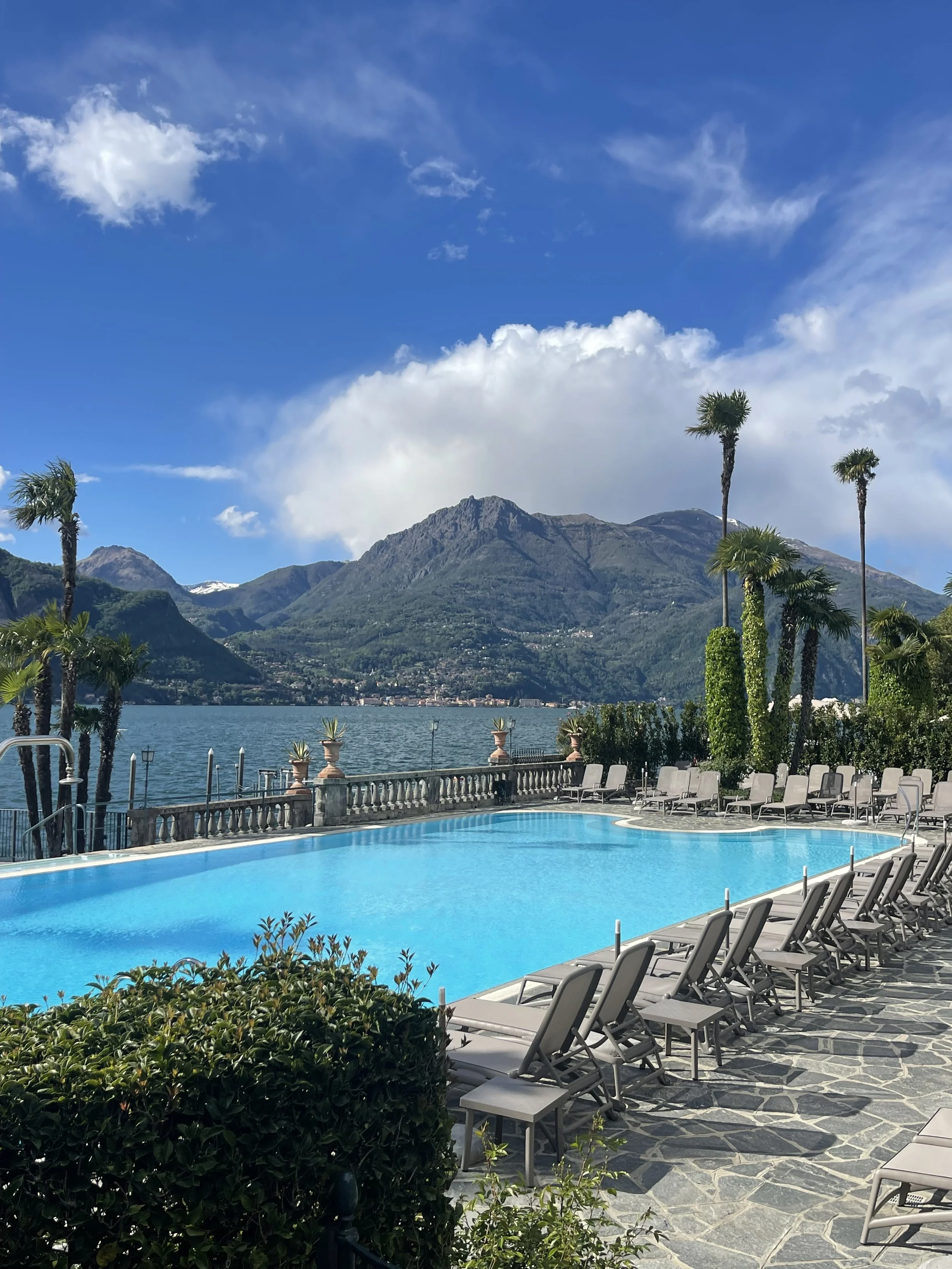 Swimming pool with lounge chairs overlooking a lake, surrounded by palm trees and mountains under a partly cloudy sky.