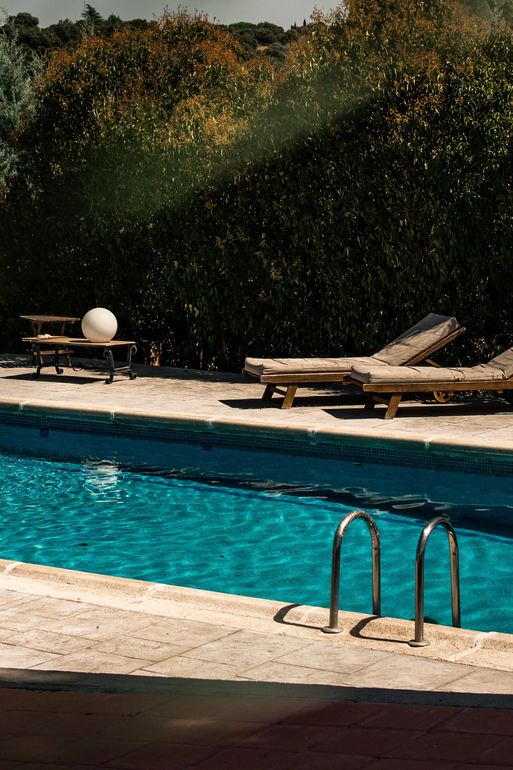 A swimming pool with clear blue water, two lounge chairs with cushions, and a small table with a decorative sphere on it. There are trees and shrubs in the background.