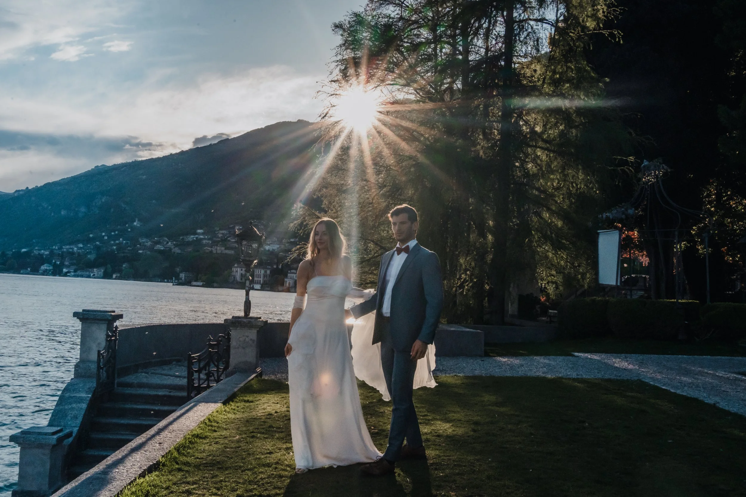 A couple in wedding attire standing outdoors by a lake at sunset, with mountains in the background and sunlight streaming through trees.