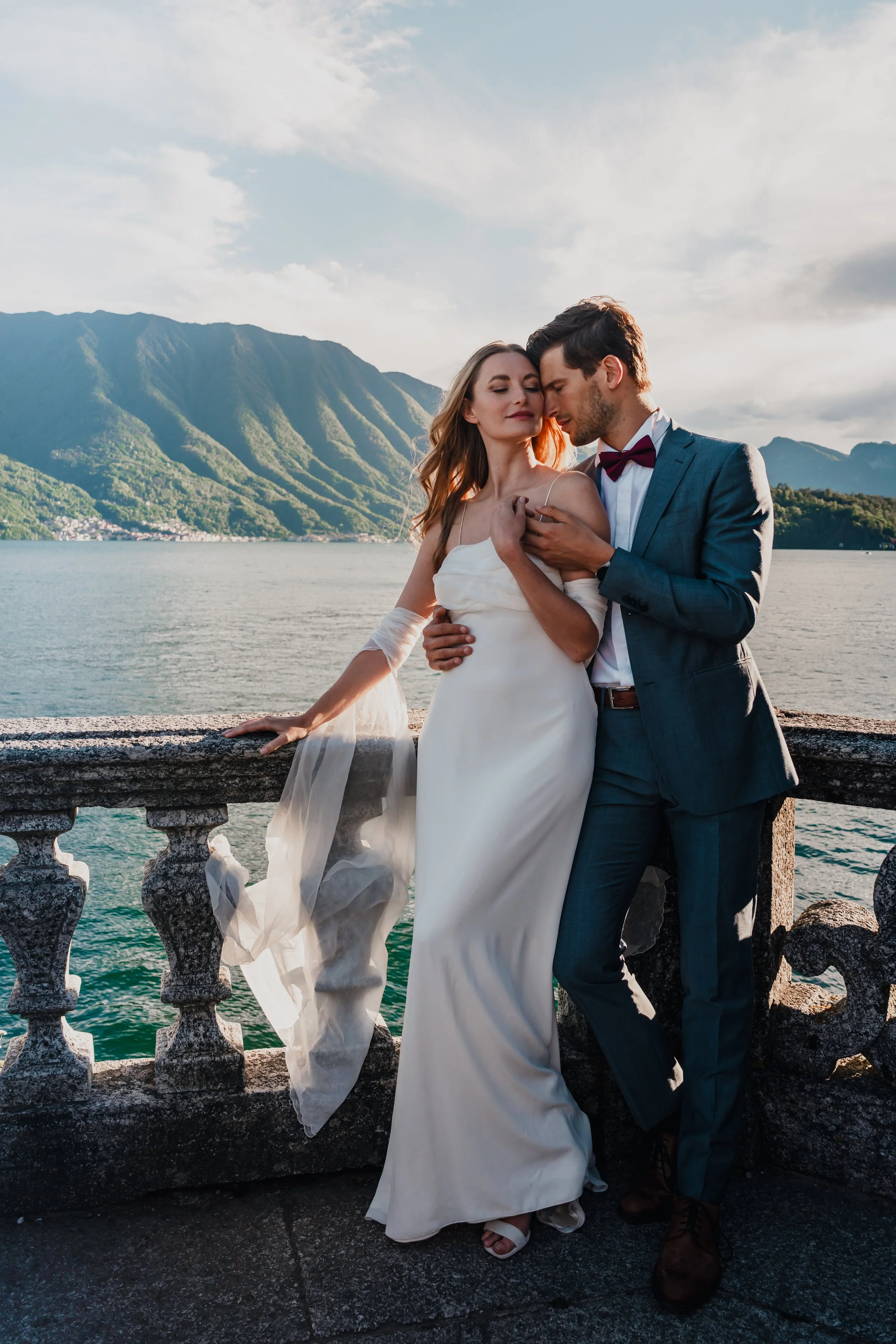 A couple in wedding attire stands on a stone railing by a scenic water body with mountains in the background, embracing each other.