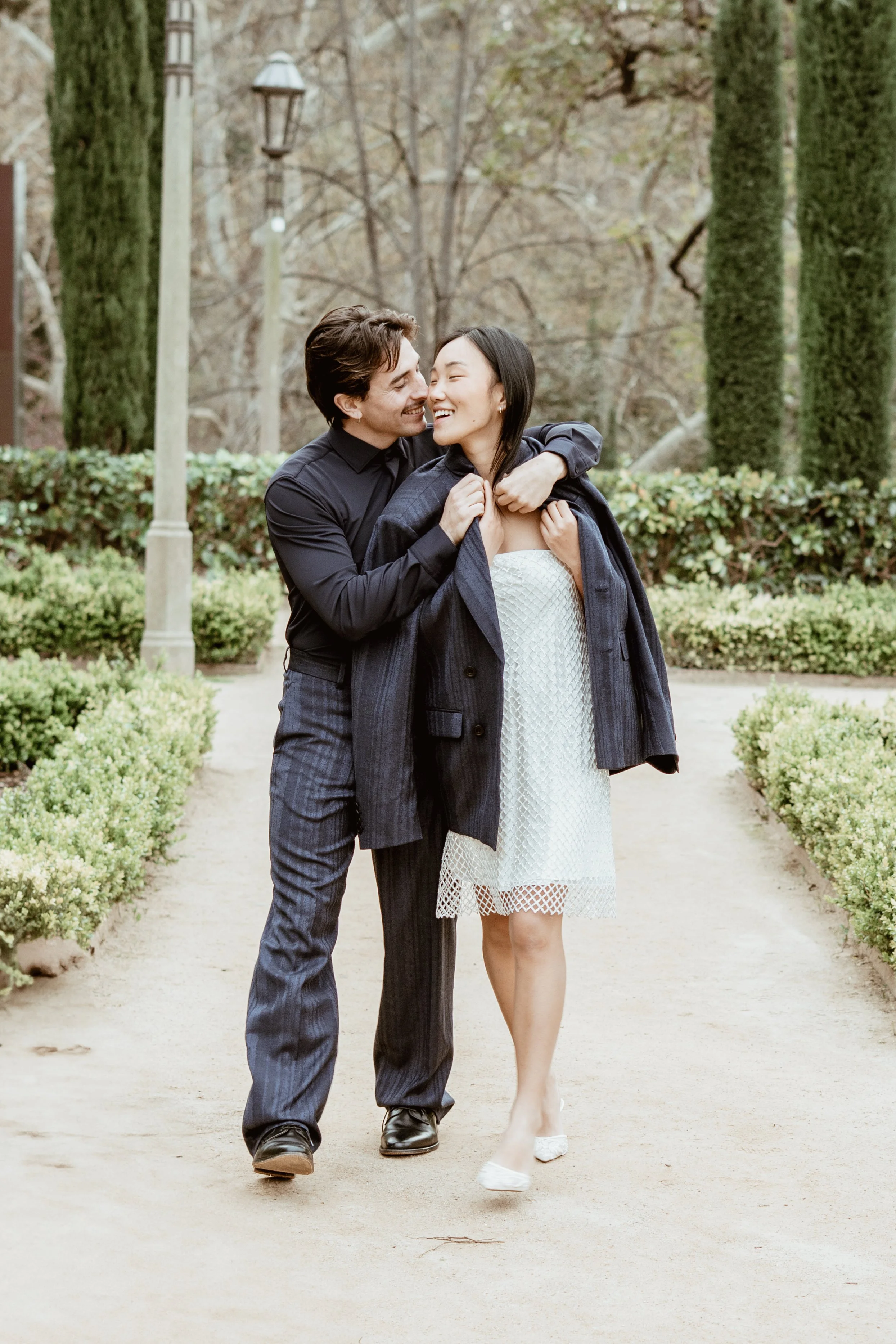 A couple walking and laughing together in a park, dressed in formal clothing, with greenery and trees in the background.