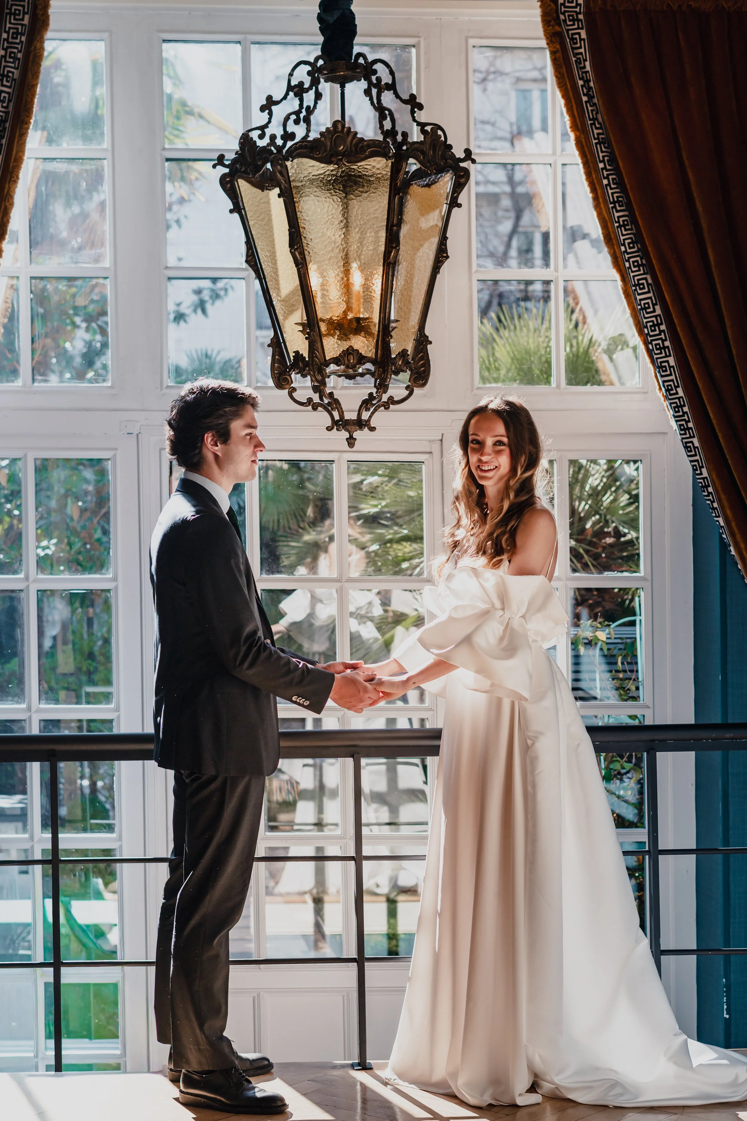 A bride and groom holding hands and smiling in front of large windows, with sunlight shining through, surrounded by elegant drapes and a chandelier hanging from the ceiling.