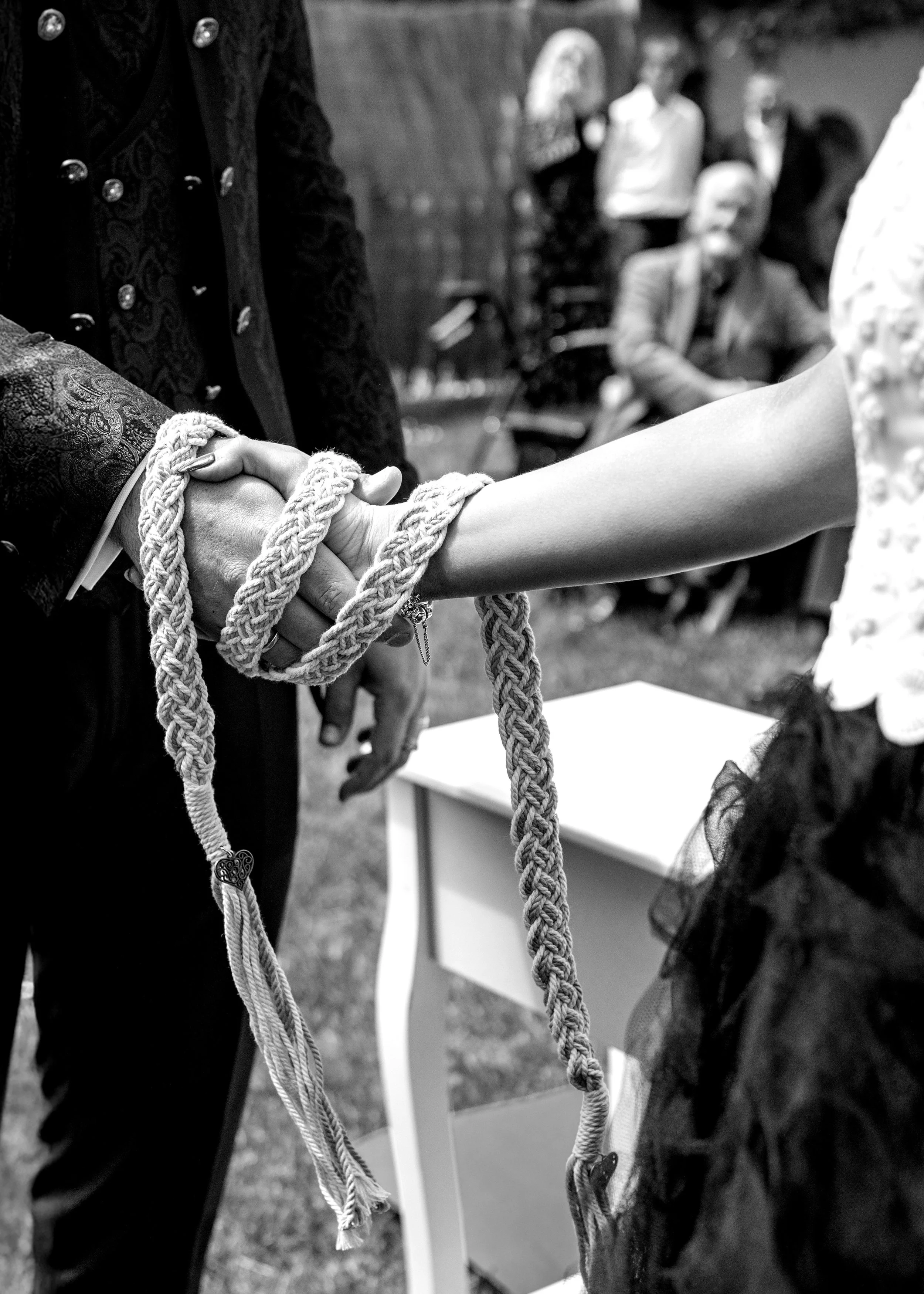 A couple exchanging rings with hands tied together by a thick rope during a wedding ceremony, with guests blurred in the background.