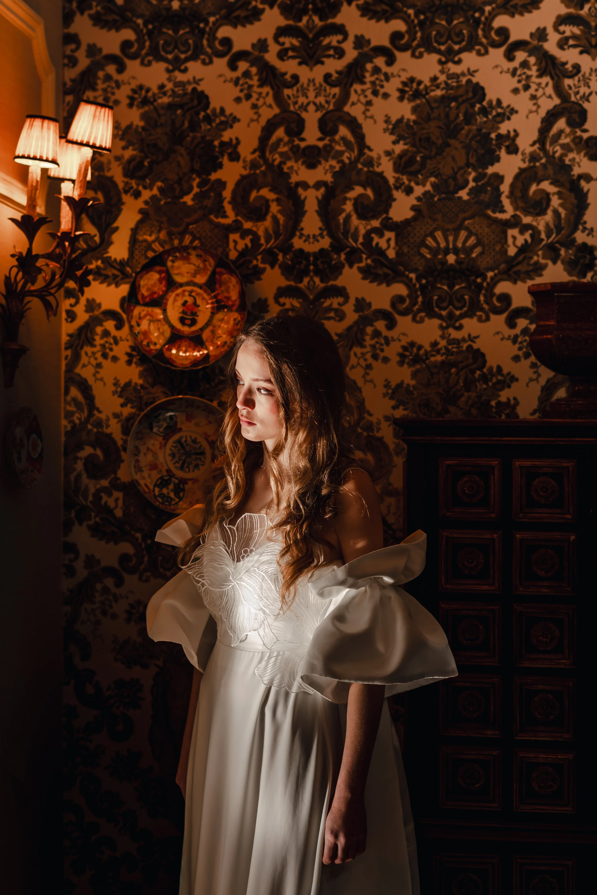 A young woman with long wavy hair in a white dress with large puffed sleeves, standing in a room with patterned wallpaper, decorative plates on the wall, and dim warm lighting.