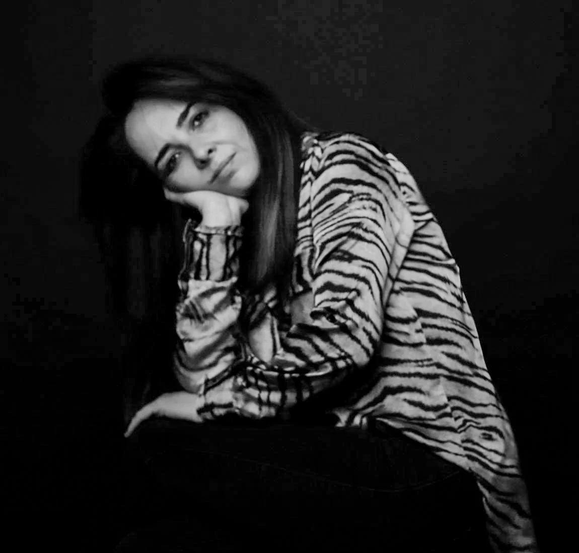 Black and white photograph of a young woman with long dark hair, resting her head on her hand, wearing a zebra print shirt, sitting against a dark background.