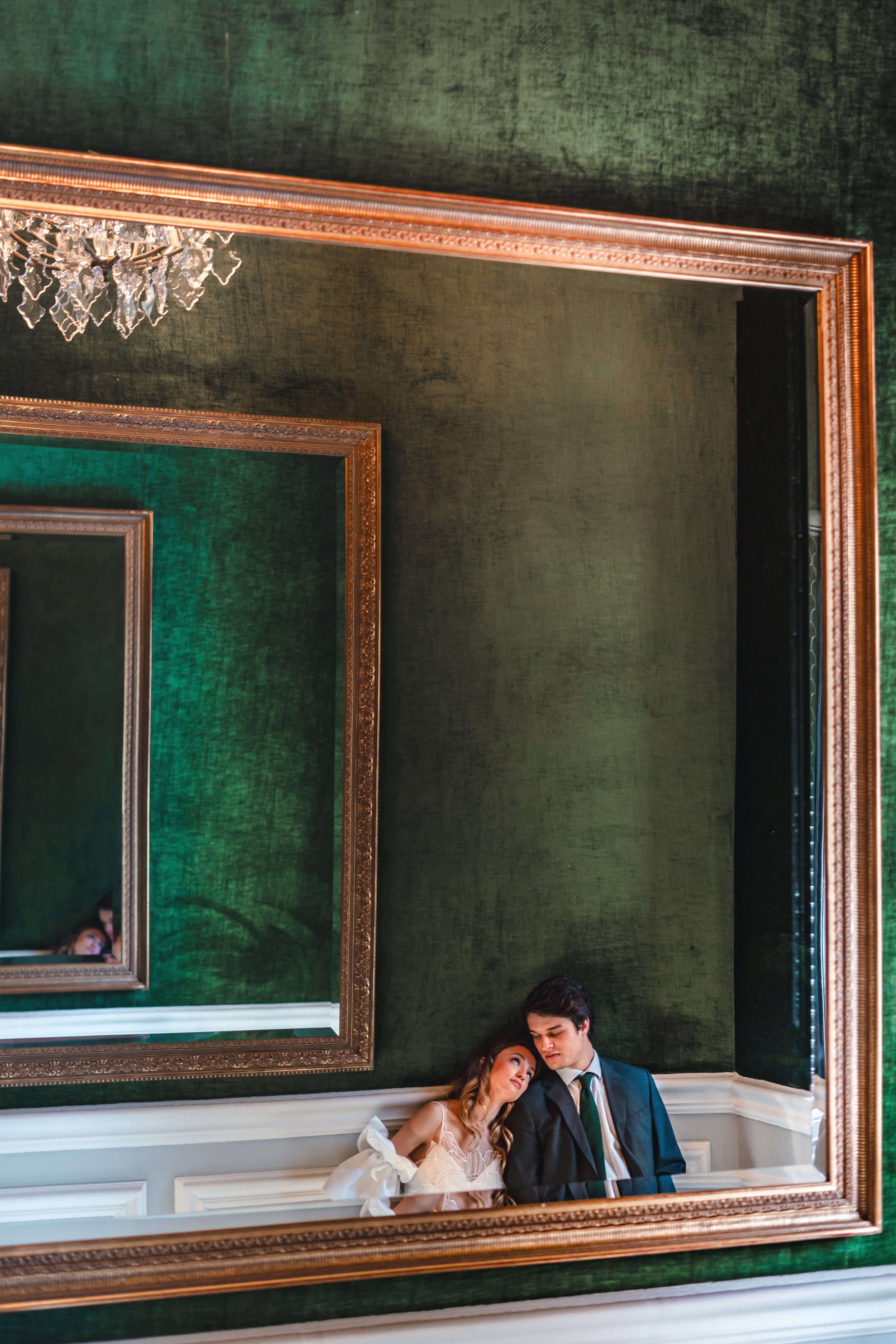A man and a woman sitting closely together, leaning their heads on each other's shoulders, reflected in a large decorative mirror with green walls and ornate gold frames.