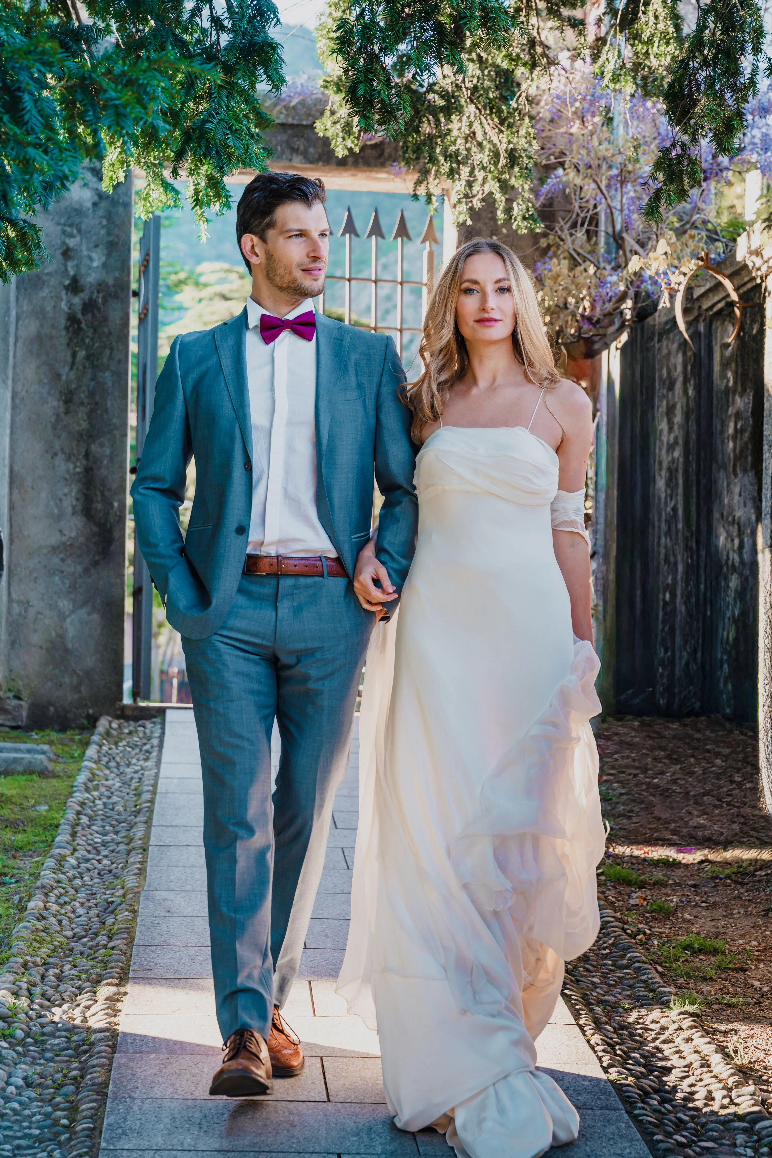 A newlywed couple walking outdoors on a sunny day, the groom in a blue suit with a purple bow tie and the bride in a white strapless wedding dress with lace gloves, under green trees.