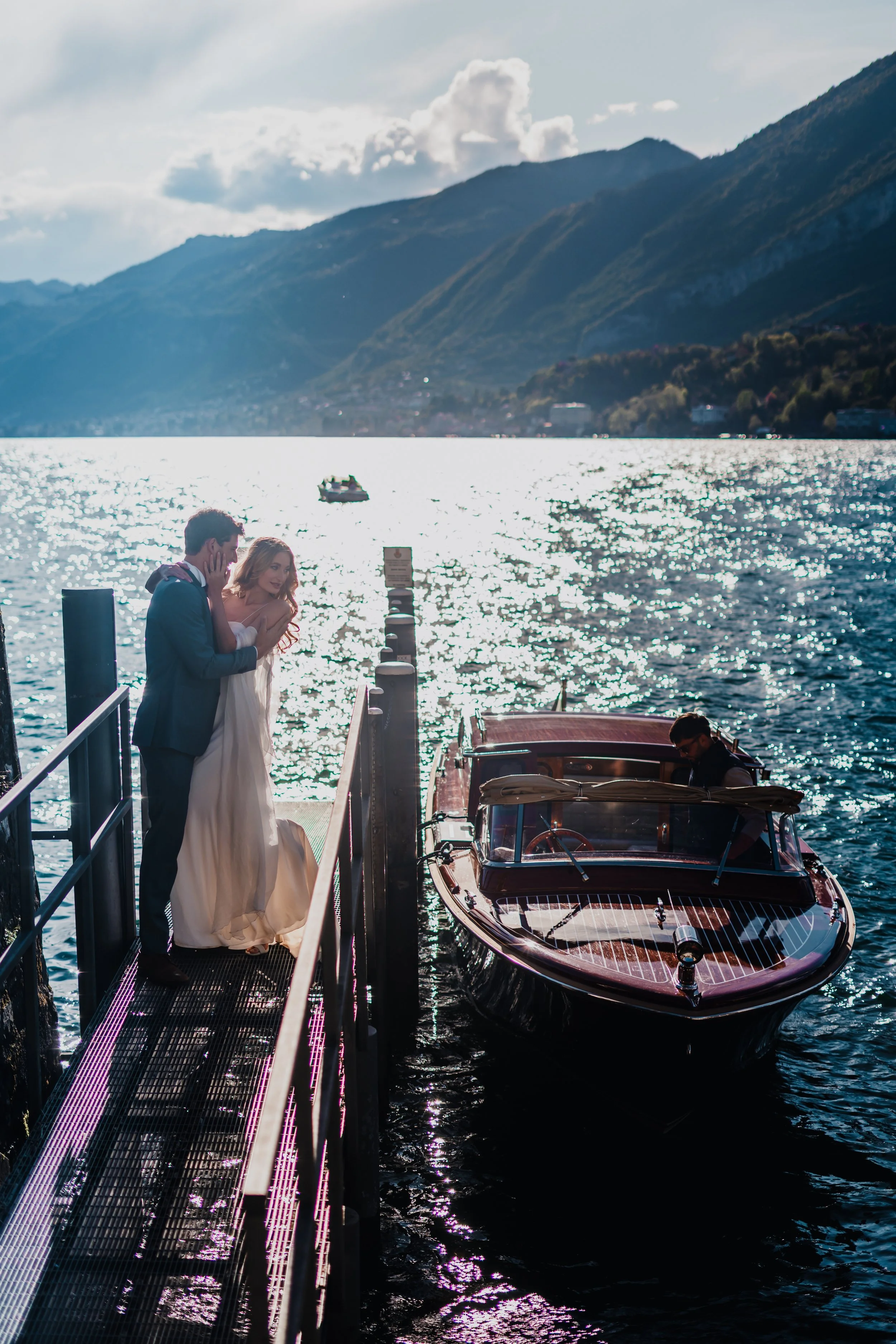 A wedding couple standing on a dock by a boat on a lake with mountains in the background during daylight.