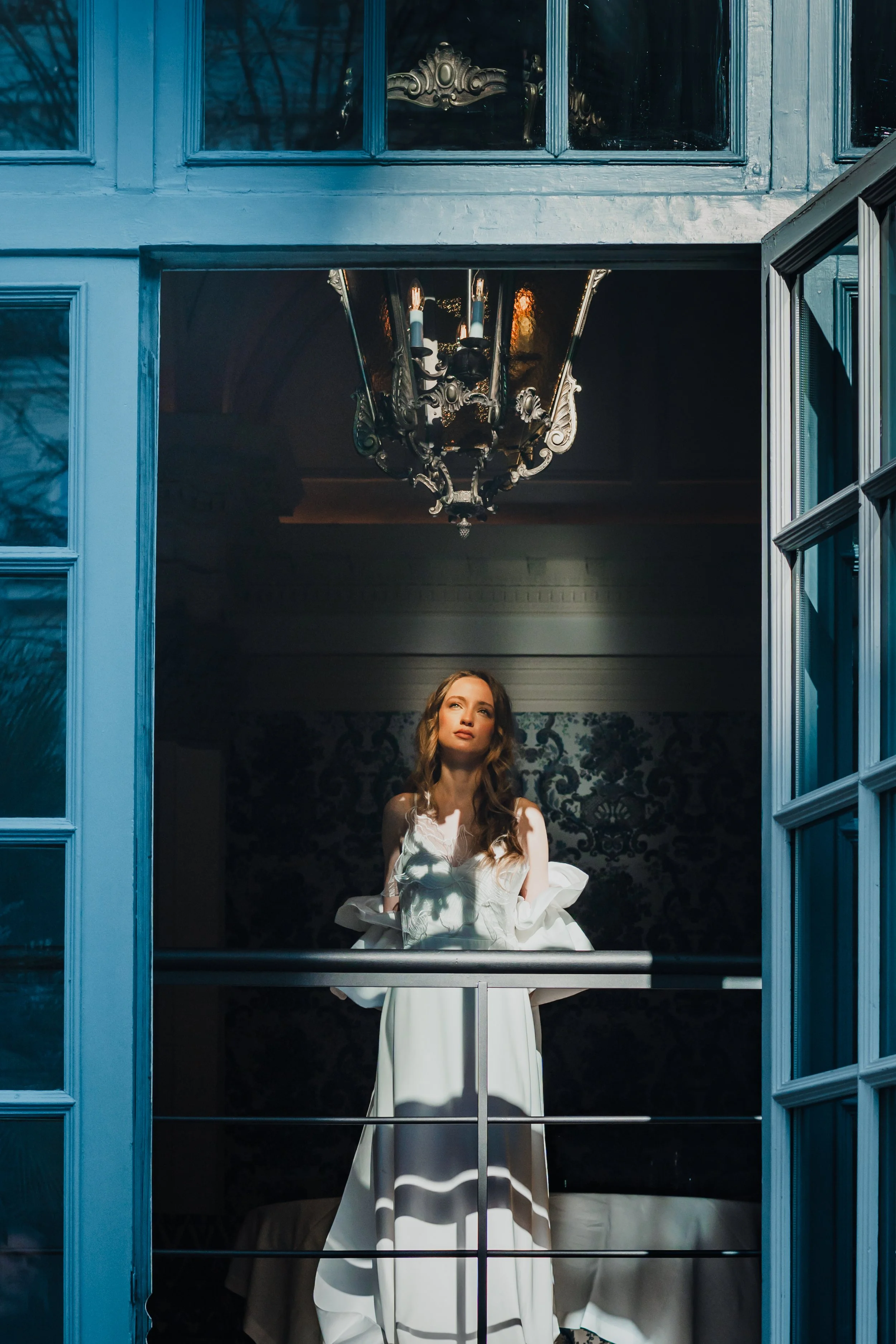 Woman in a white dress standing on a balcony indoors, looking up, with a chandelier hanging above her and blue window frames surrounding her.