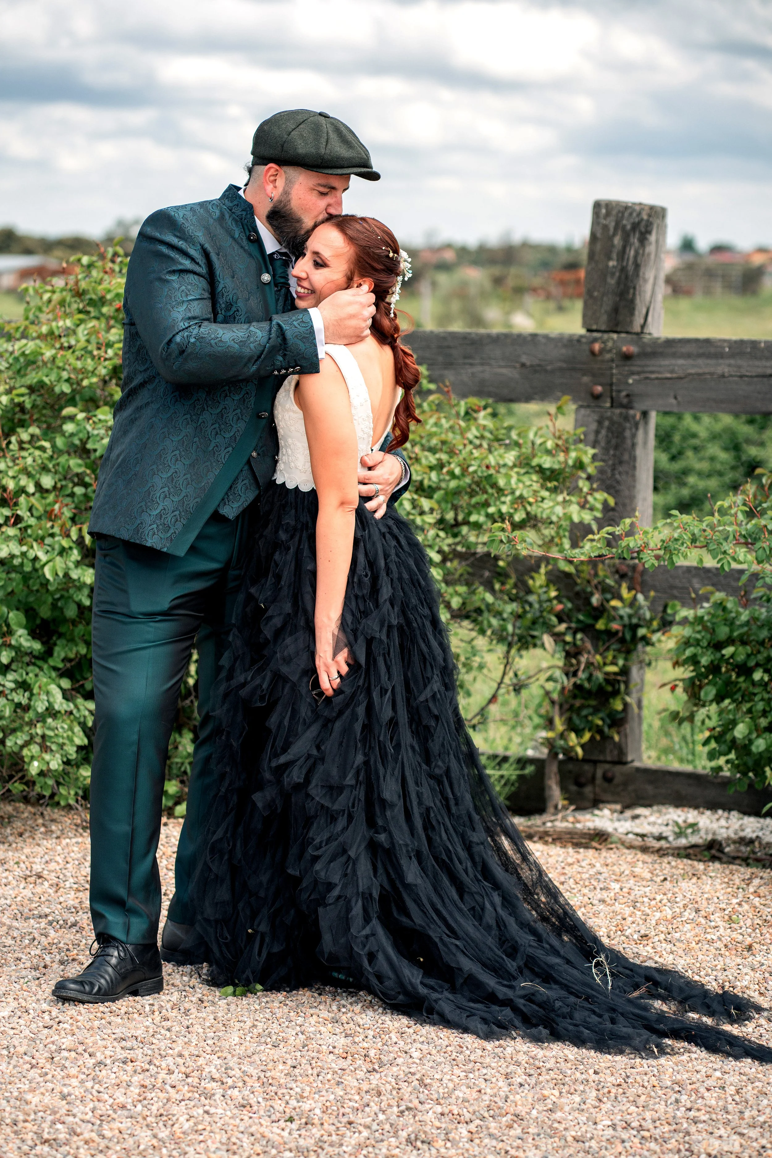 A couple on their wedding day, with the man kissing the woman's forehead, outdoors with greenery and a wooden fence in the background.