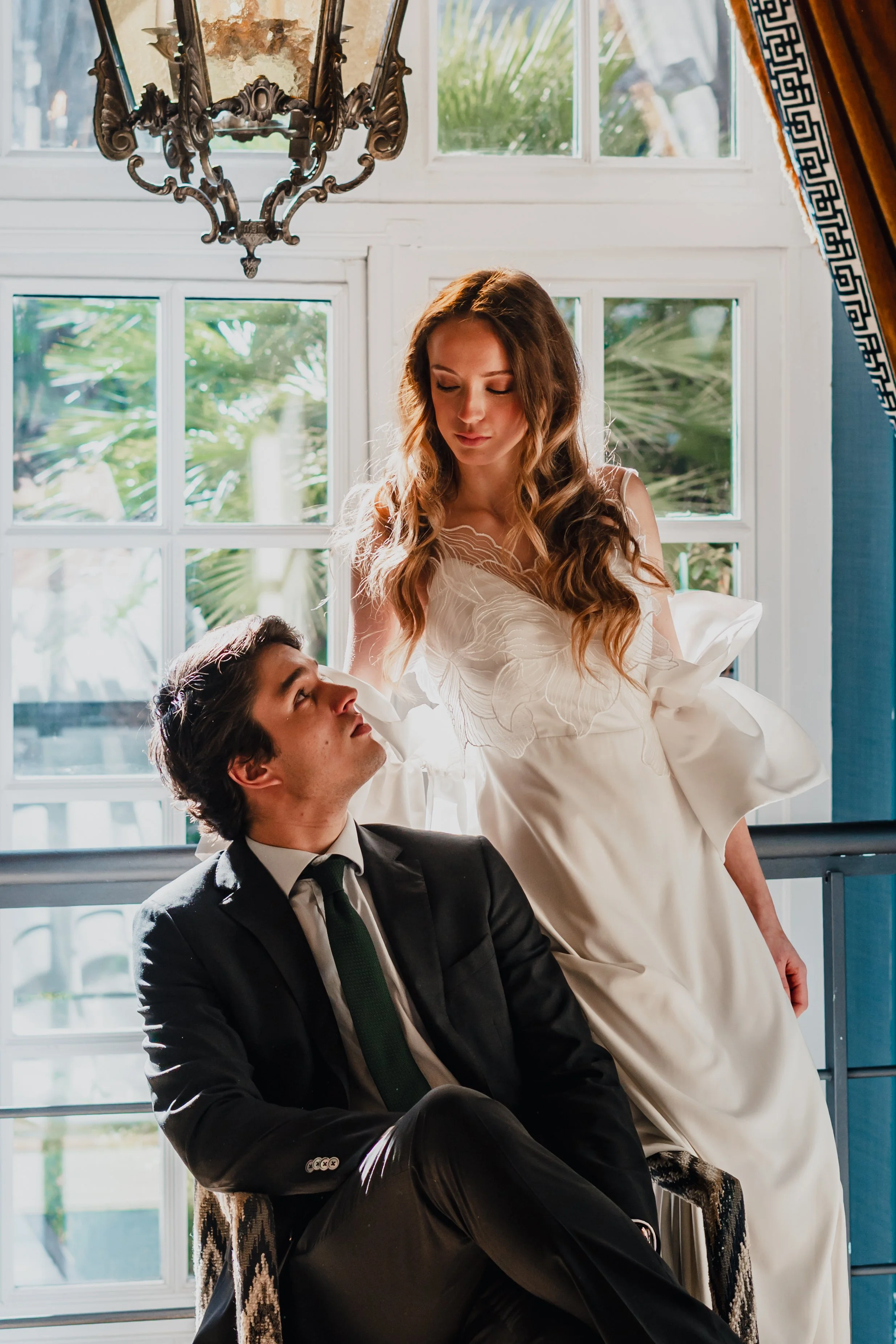 A woman in a white dress leaning towards a seated man in a black suit, inside a bright room with large windows and plants outside.