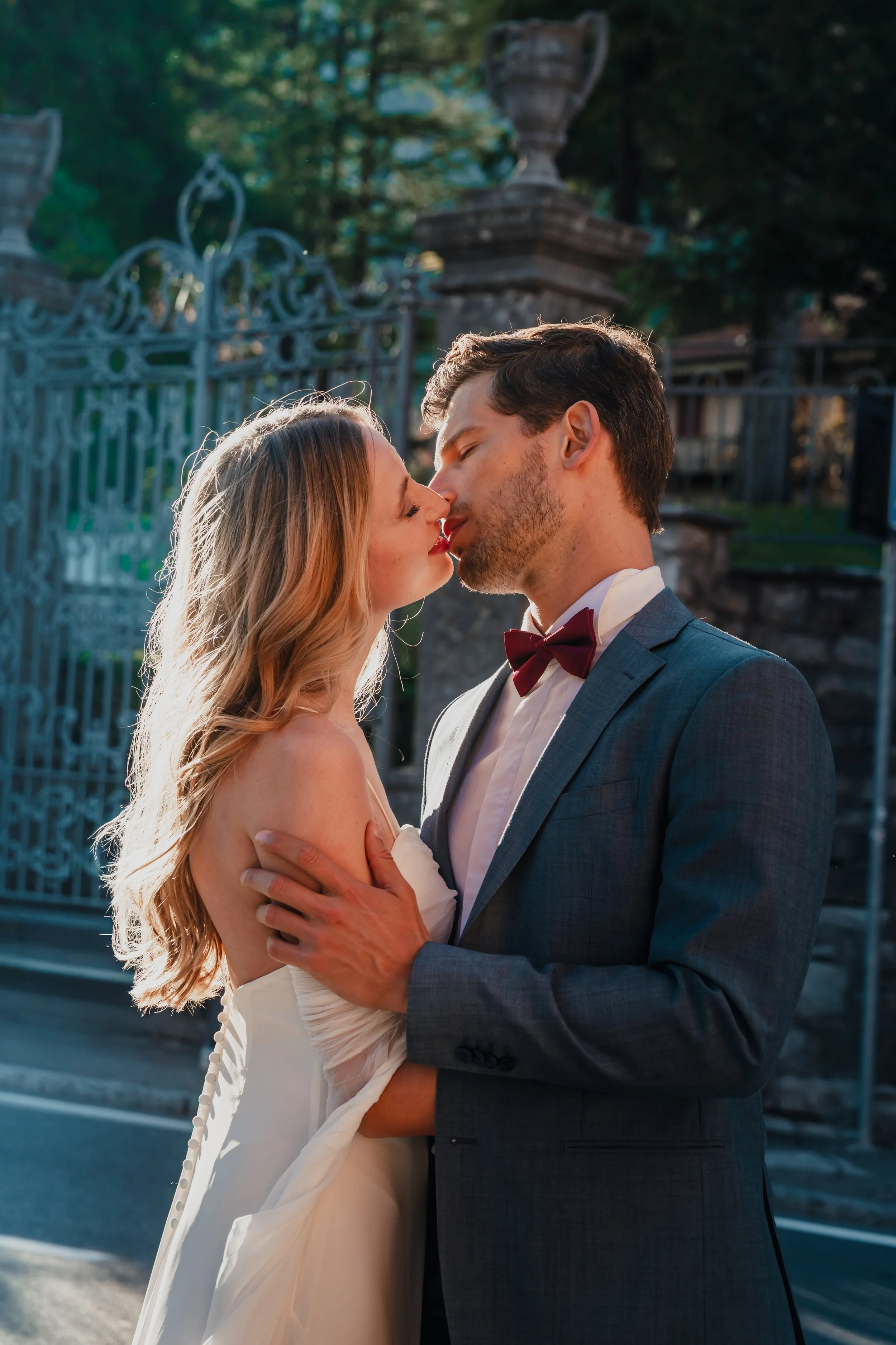 A bride and groom sharing a kiss outdoors, dressed in wedding attire, with a decorative metal gate and trees in the background.