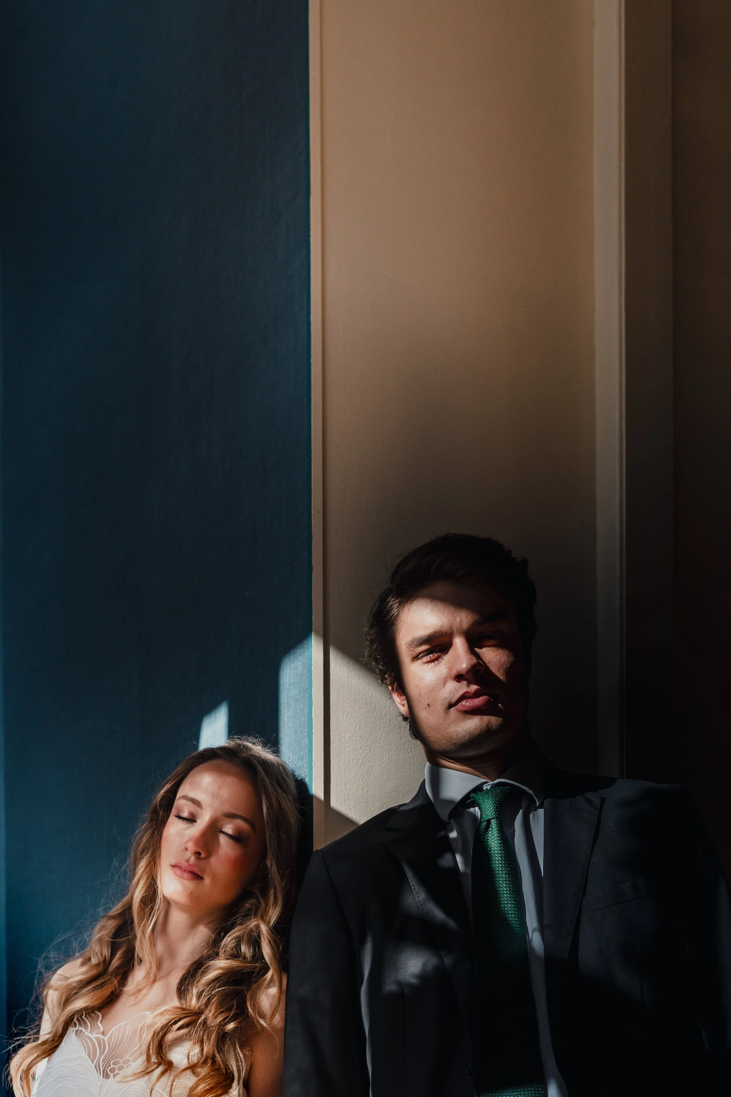 A man and woman in formal attire standing in a room with sunlight casting shadows on their faces.