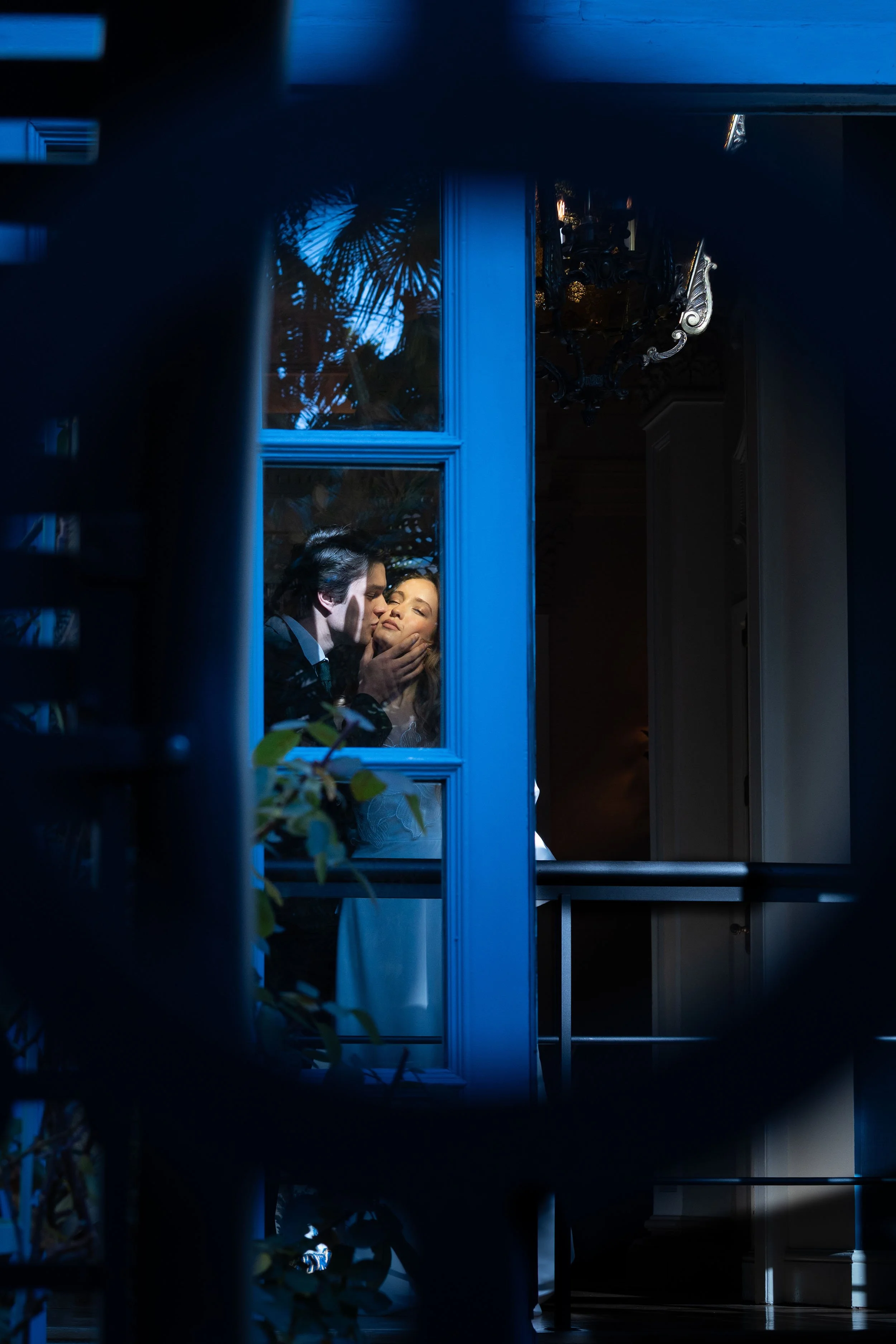 Una pareja en un momento romántico, él la besa en la frente dentro de una habitación visible a través de una ventana azul