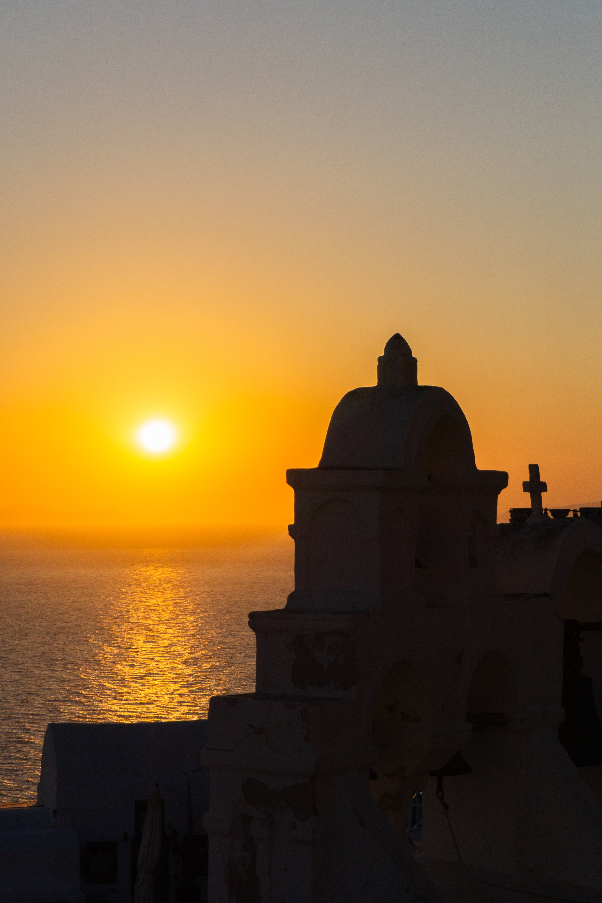 Sunset over the ocean with a silhouette of a church or building with a small dome and cross in the foreground.
