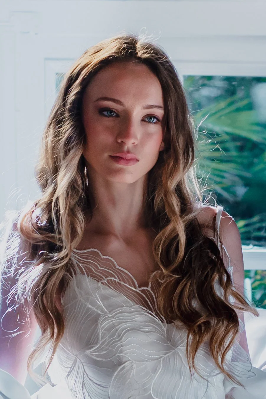 A woman with long, wavy brown hair and blue eyes looking at the camera inside a bright room with a window and green plants in the background.