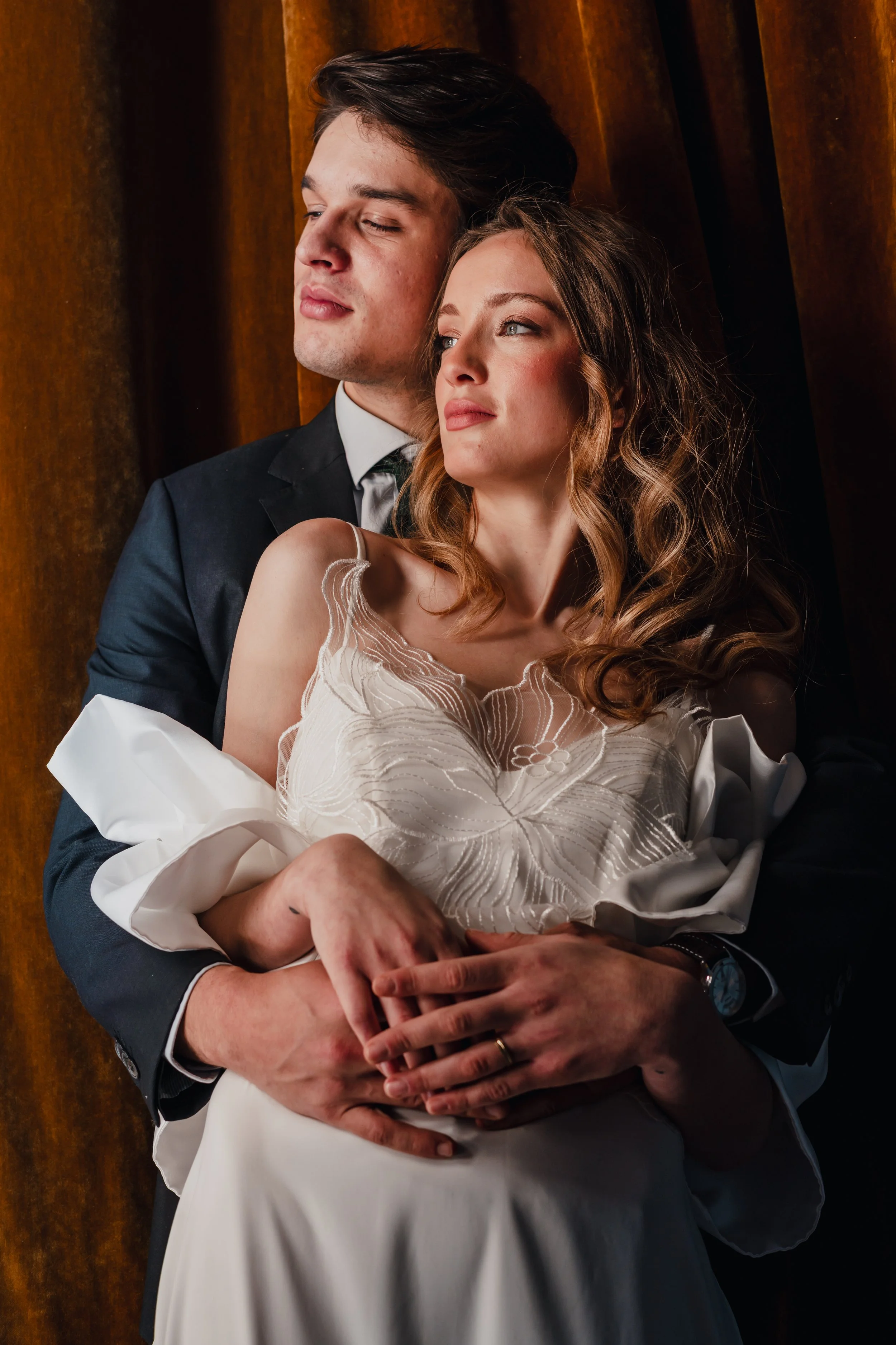 A man and woman in formal attire, embracing, with the man holding the woman from behind, and they are sitting against a wooden background.