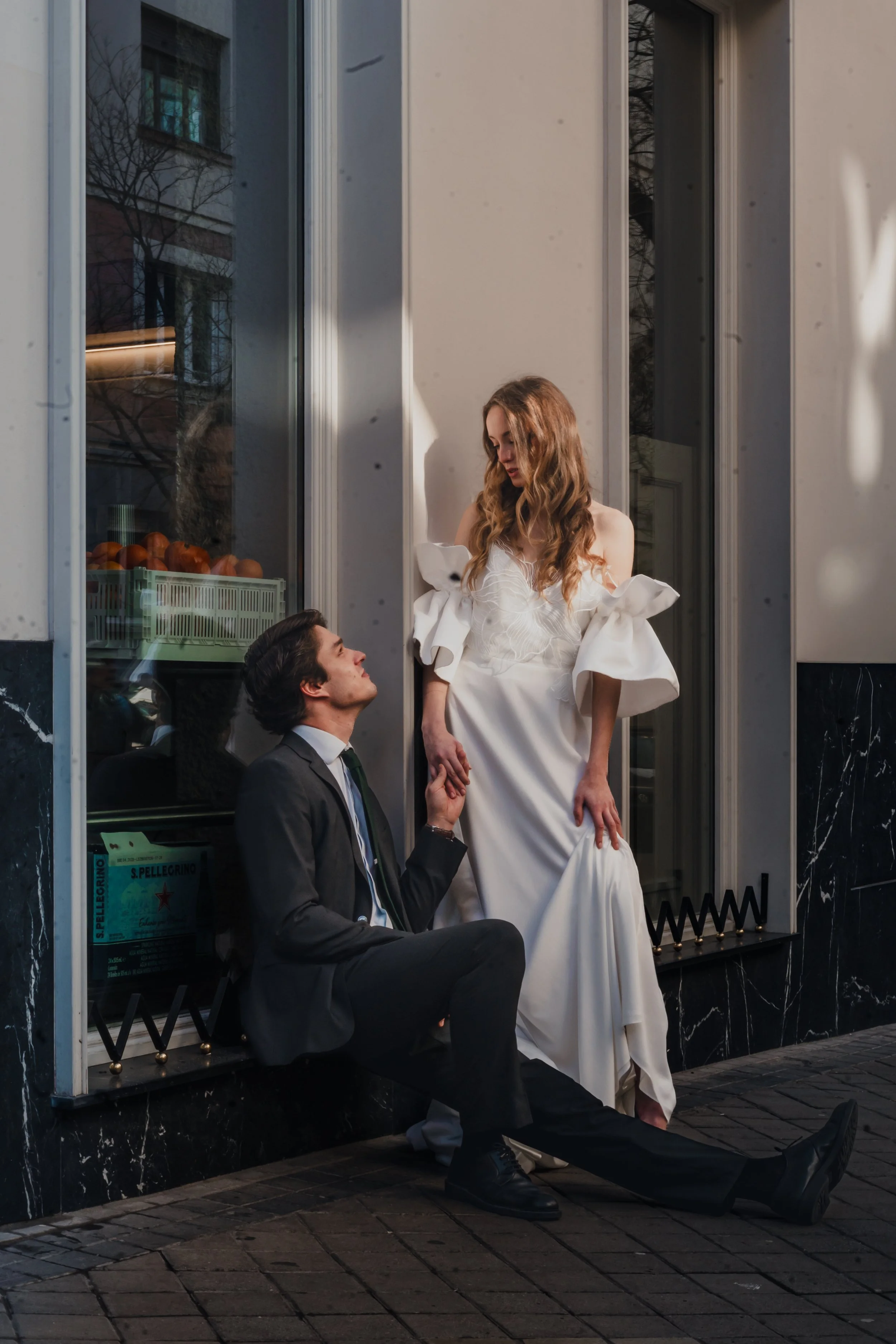 A man dressed in a suit sitting on the sidewalk, holding hands with a woman in a white dress standing next to him, outside a building with large windows.