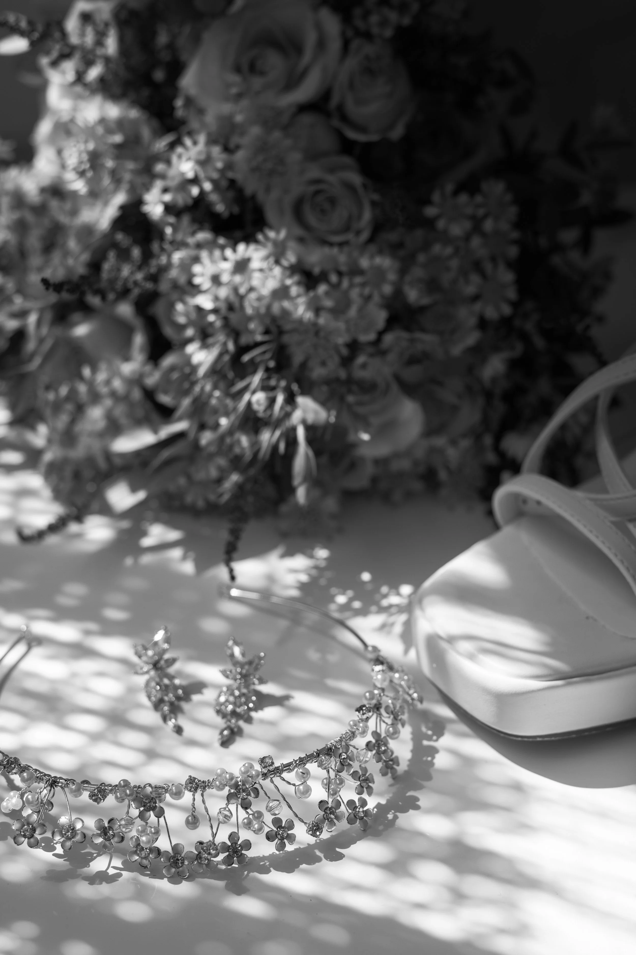 A wedding bouquet, a tiara, and a white high-heeled shoe on a table, with floral and shadow patterns.