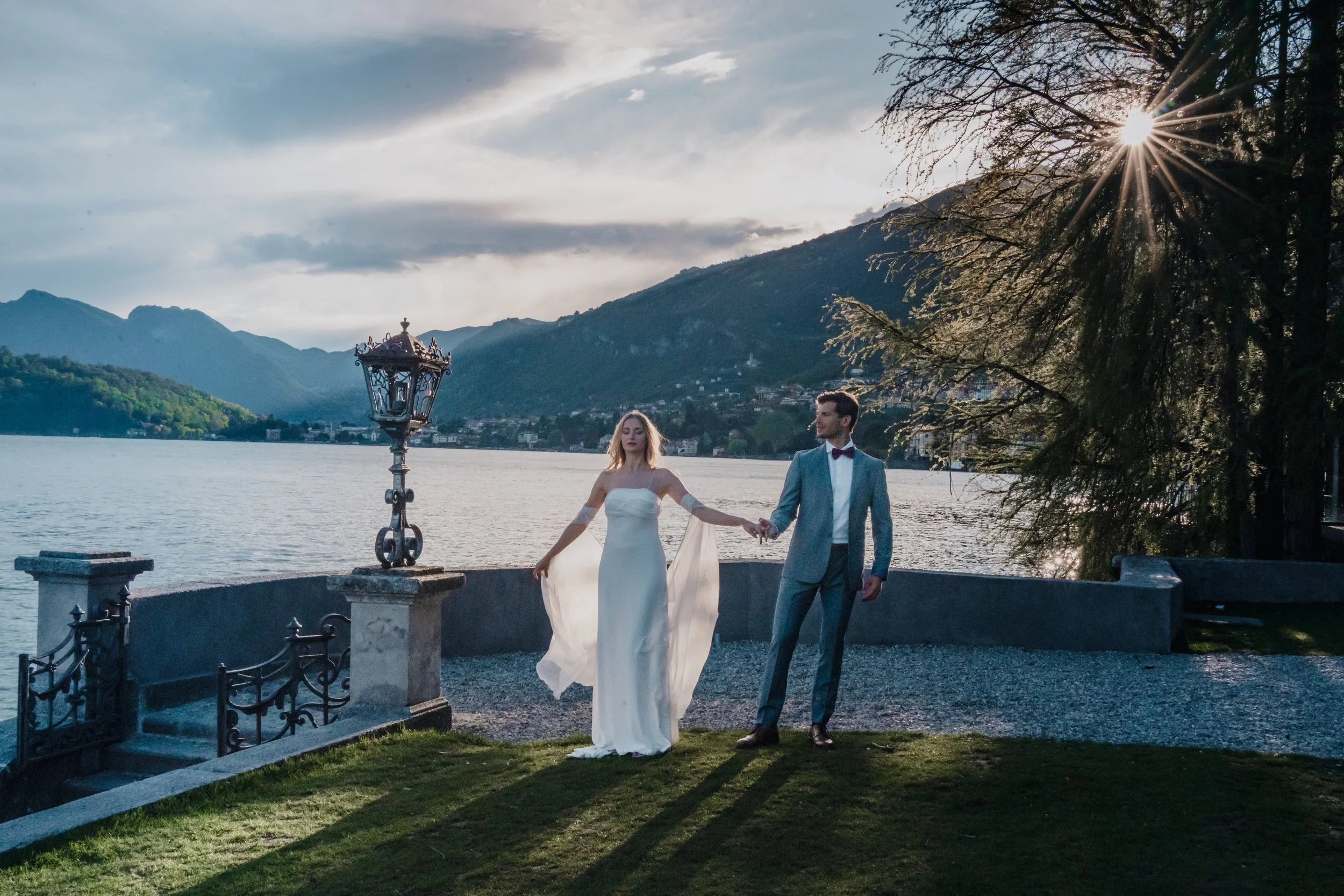 A bride and groom holding hands by a lake during sunset, with mountains in the background and a tree partially blocking the sun.