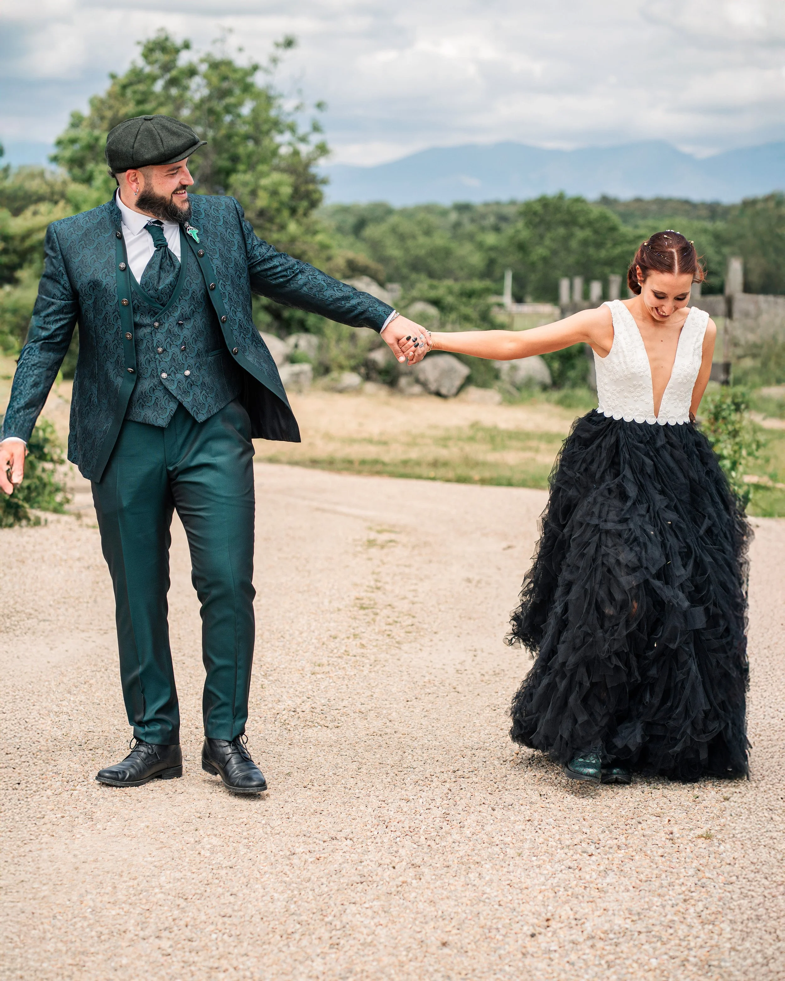 A man in a teal suit holding hands with a woman in a black and white gown, walking on a dirt path outdoors.