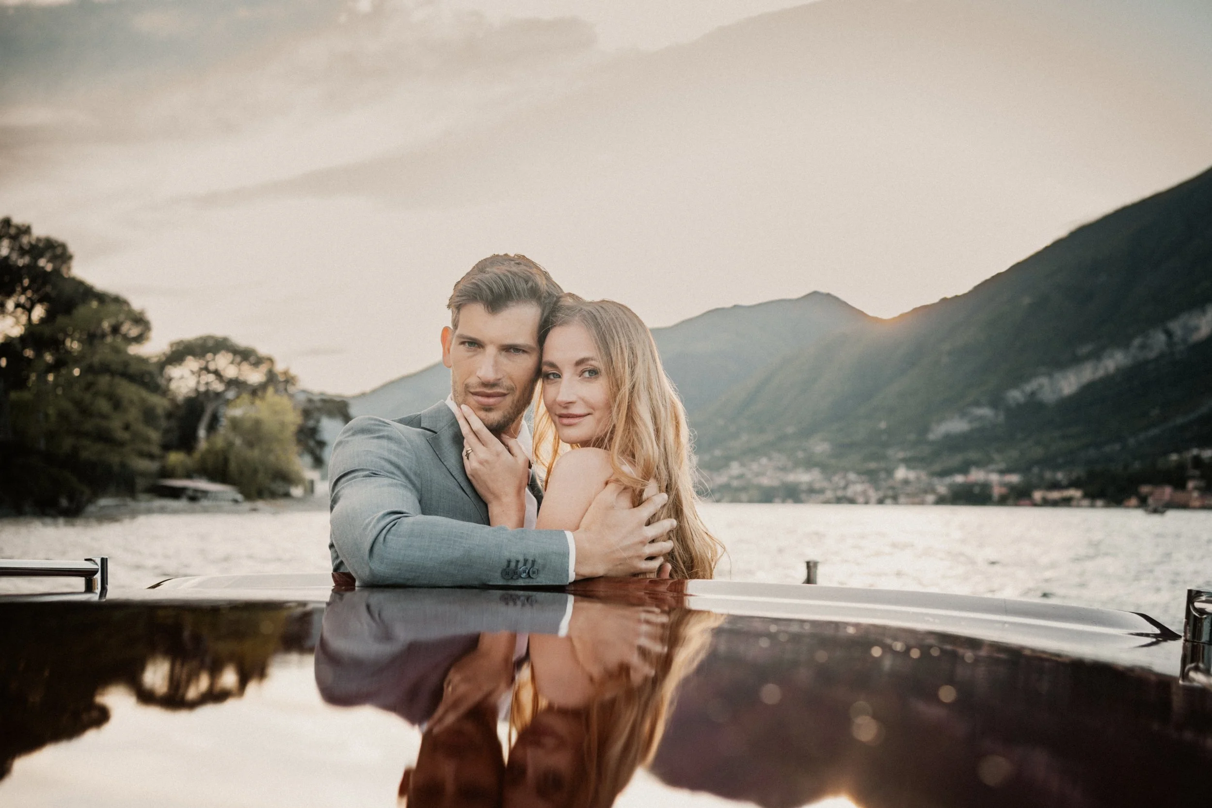 A couple embracing on a boat with a lake and mountains in the background during sunset.