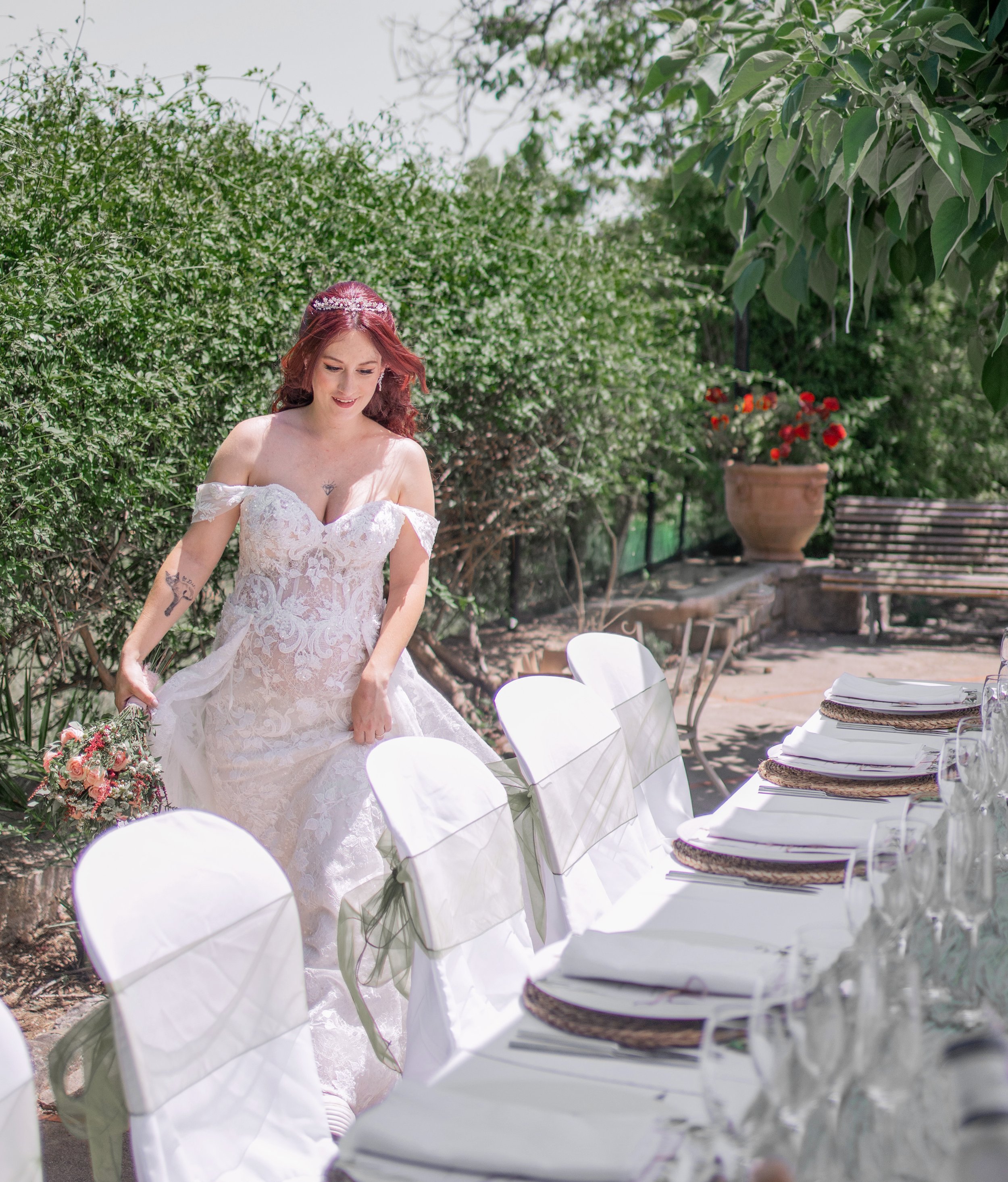 A bride in a white lace wedding dress with off-the-shoulder sleeves holding a bouquet of pink and white flowers, standing outdoors near a table set for a wedding reception.