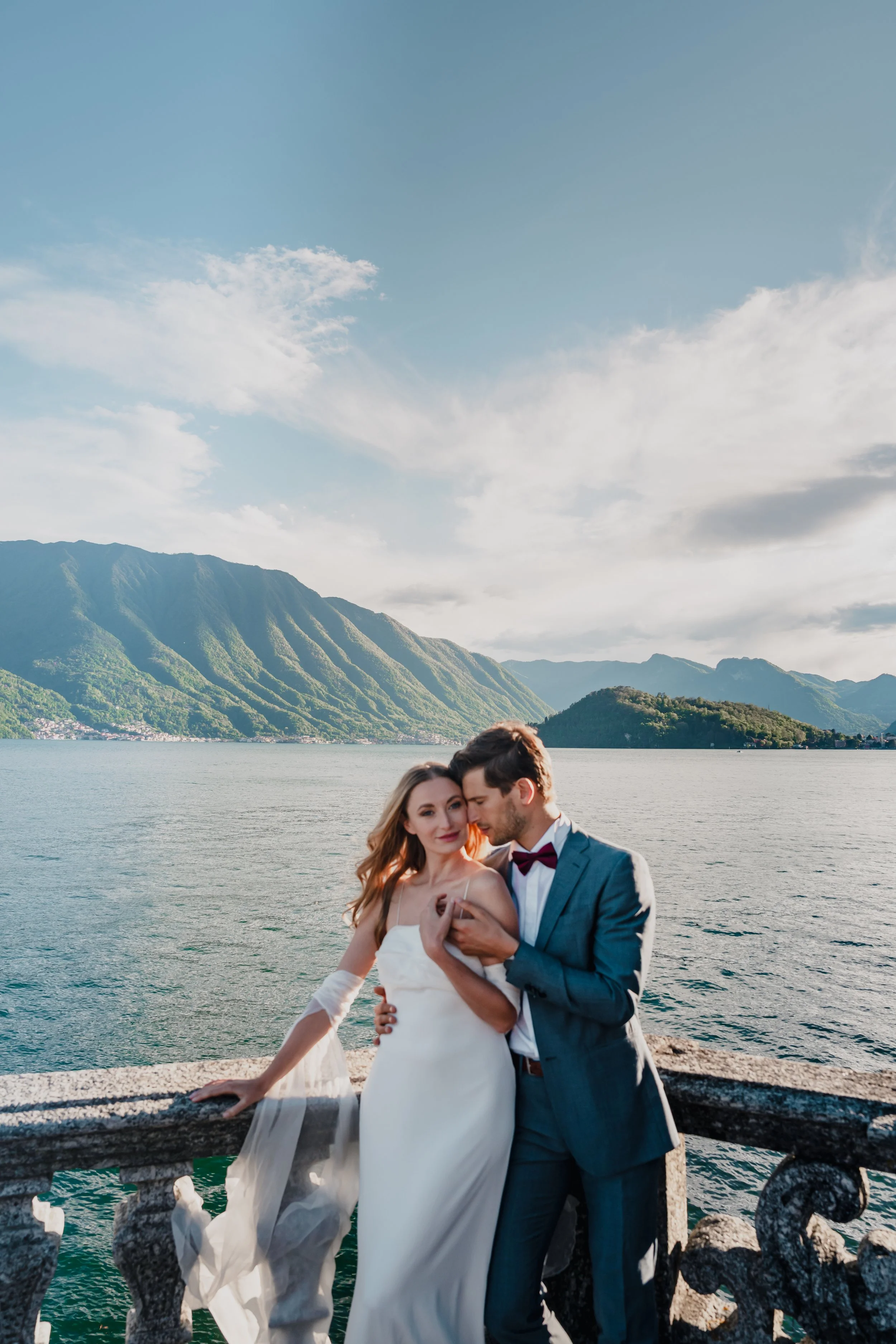 A bride and groom standing by a lake with mountains in the background. The bride is wearing a white wedding dress and the groom a suit with a bow tie. They are embracing each other on a stone railing.
