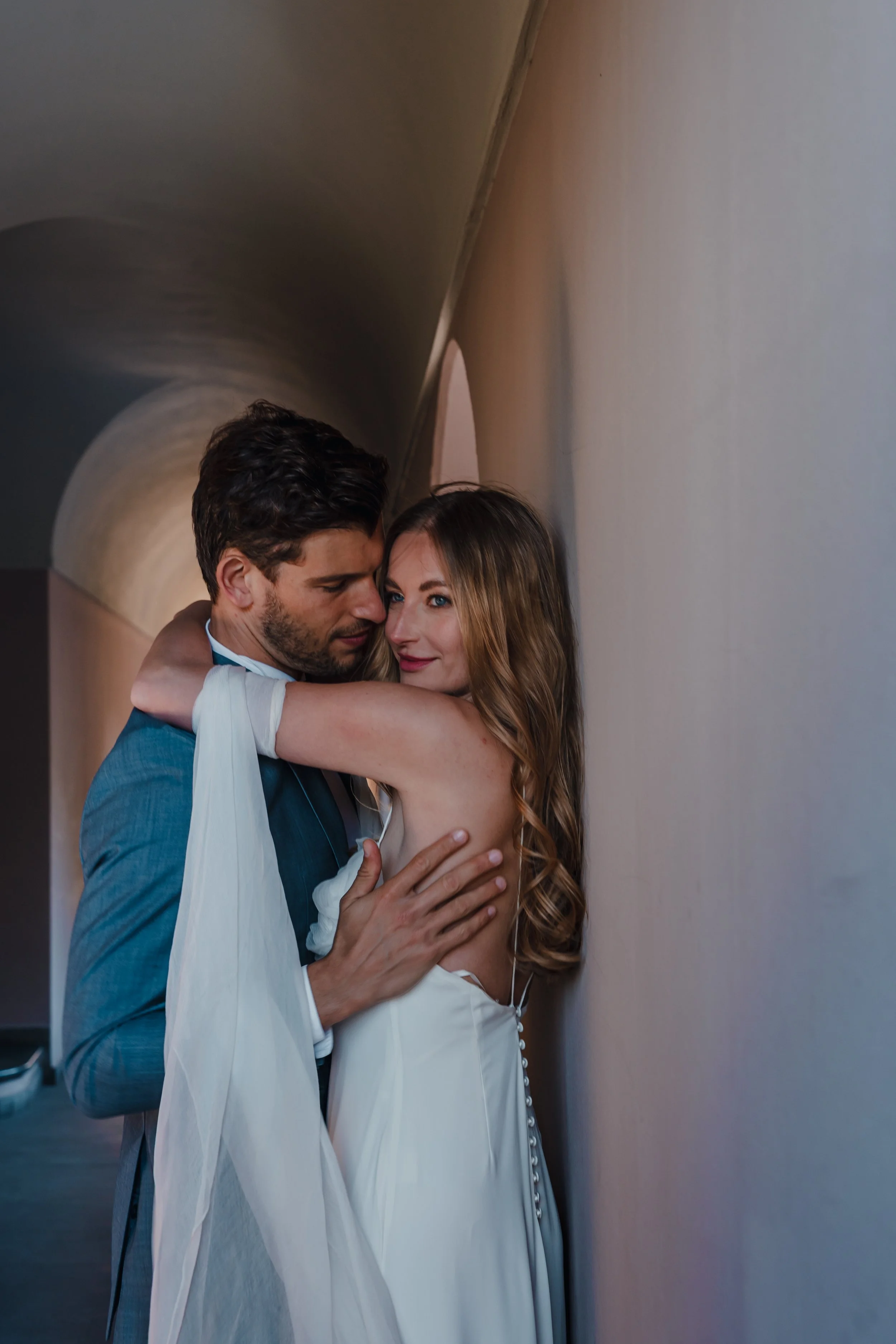 A couple dressed in wedding attire hugging against a curved wall at an indoor venue.